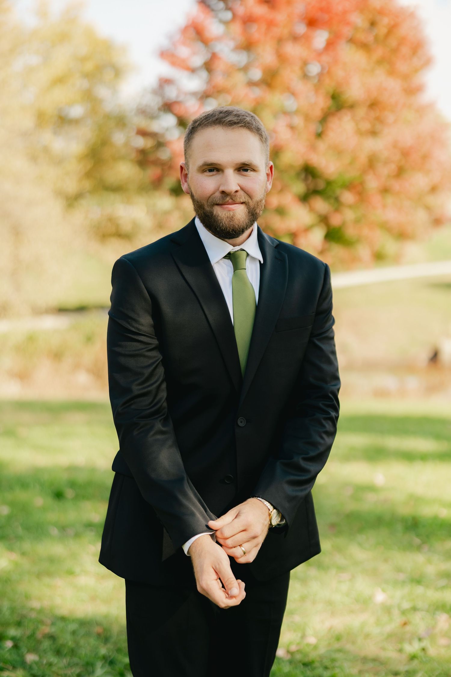 Bearded man in a black suit, white shirt and green tie adjusts his cuff while smiling outdoors on a lawn with vibrant autumn trees.