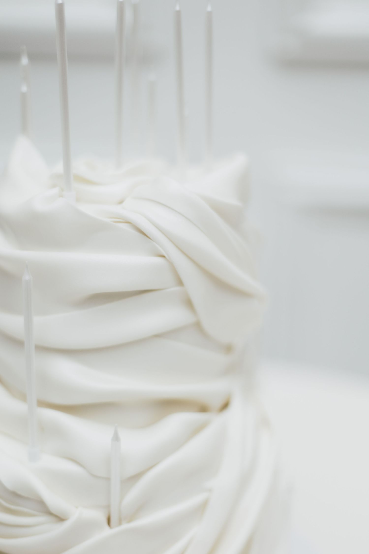 Elegant white wedding cake with draped fondant ruffles and tall white candles on a white background.