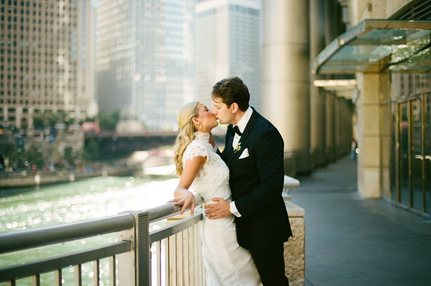 Bride and groom share a romantic kiss on a city riverwalk with Chicago skyline and river in the background.