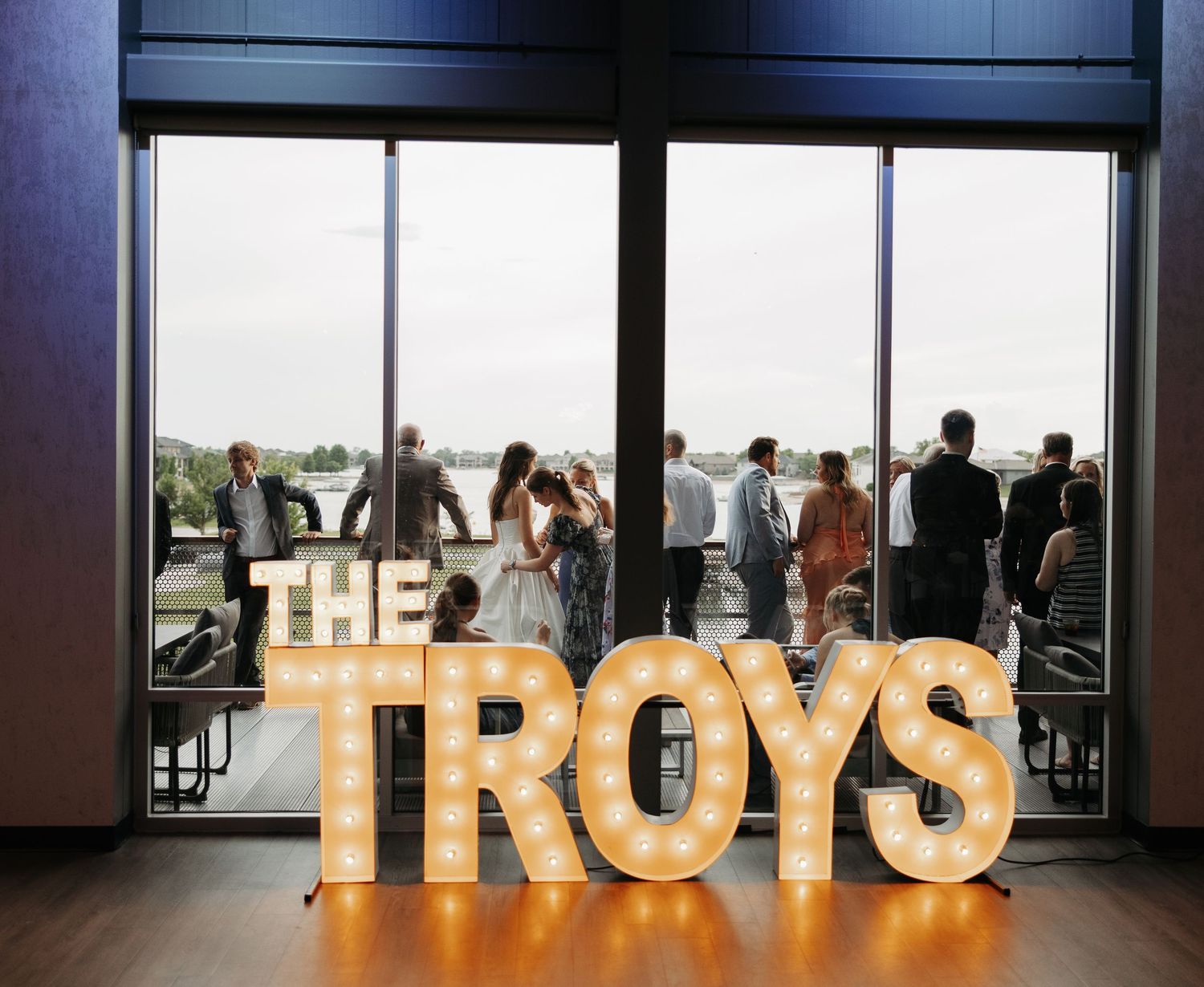 Illuminated marquee letters spelling THE TROYS at a wedding reception with guests gathered by large windows.