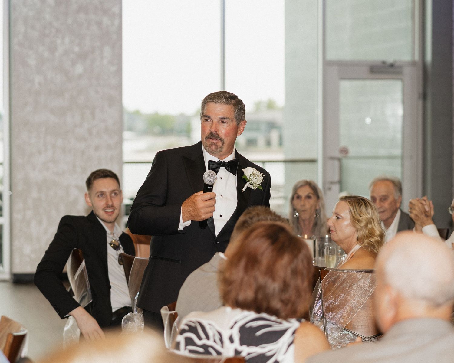 Man in tuxedo giving a wedding speech at a reception while guests listen attentively in an elegant venue.