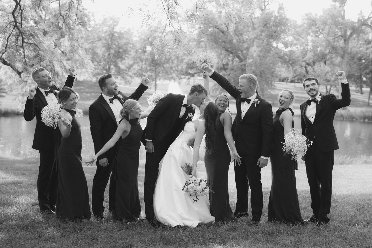 Wedding party celebrating outdoors, bride and groom kissing while guests raise glasses near a pond.