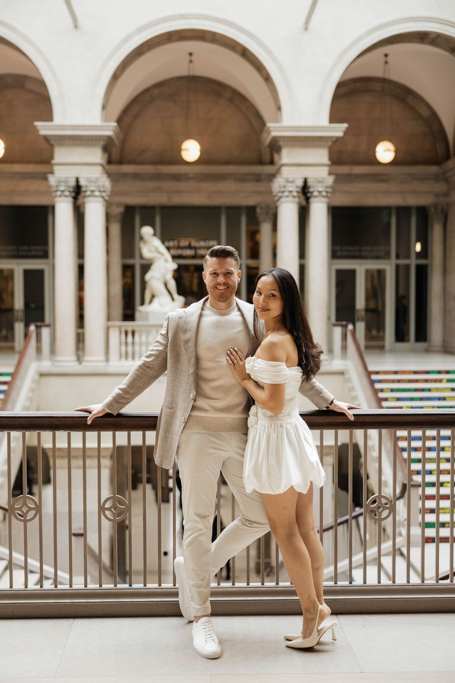 Elegant couple posing in white outfits on a balcony inside a grand museum atrium with arched ceilings.