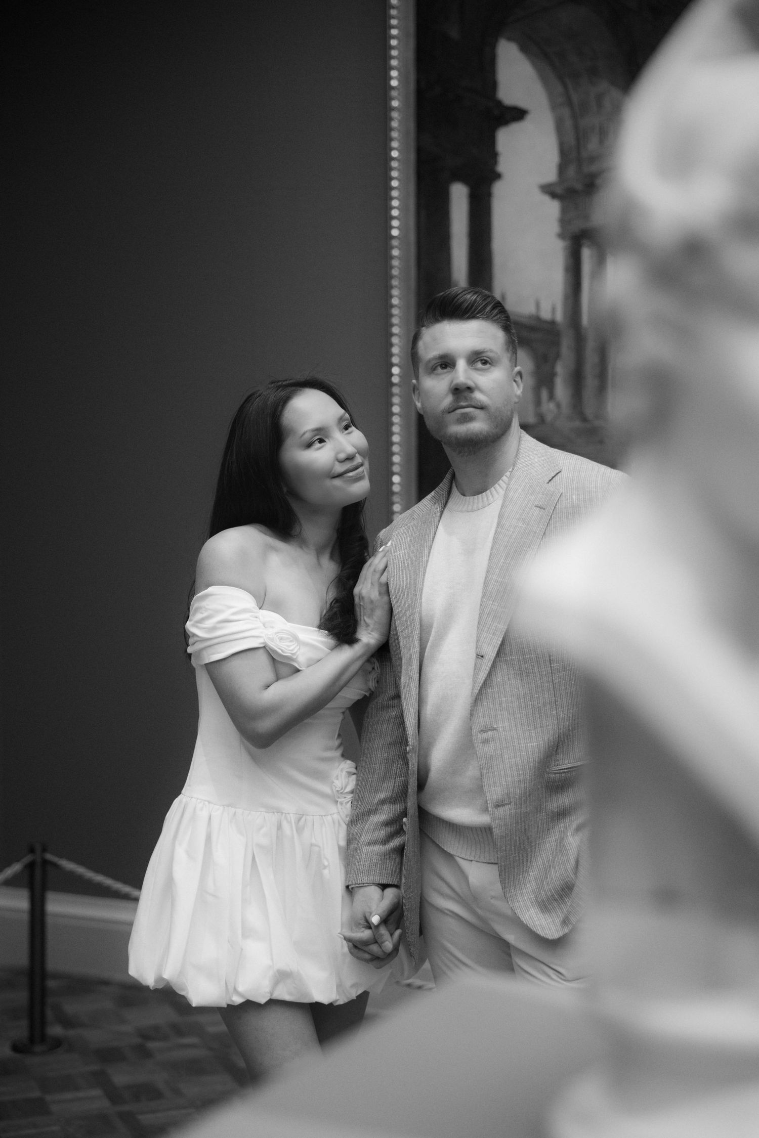 Black and white photo of a couple holding hands in an art museum, woman in white dress leaning on man in blazer.