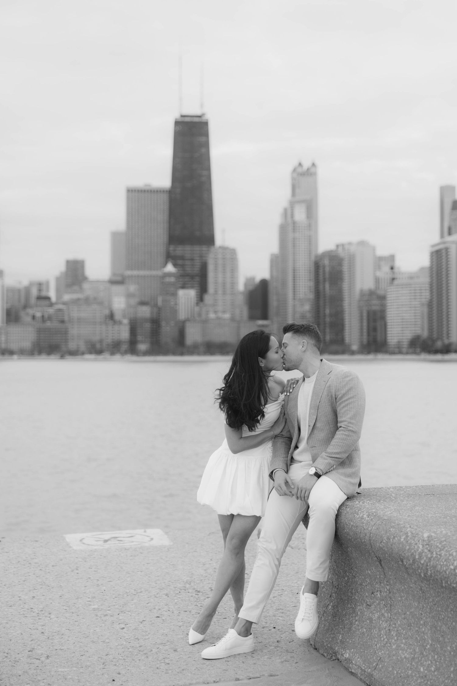 Black and white photo of couple kissing by Lake Michigan with Chicago skyline in background.