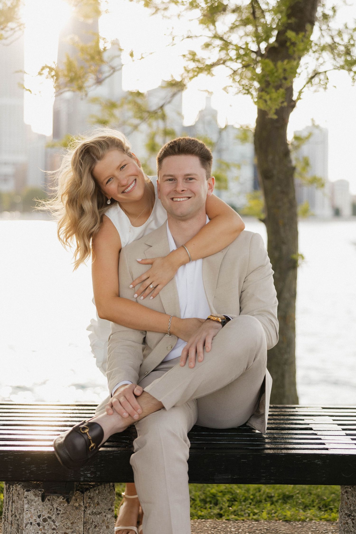 Smiling couple posing on a park bench by the water, woman embracing man from behind.