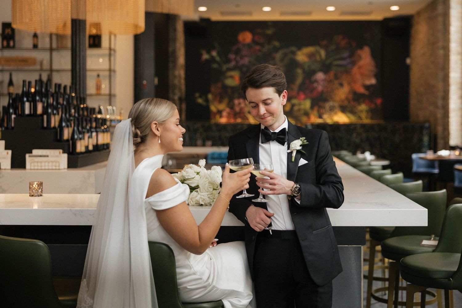 Bride and groom sharing a toast at elegant bar with wine bottles and floral artwork backdrop.