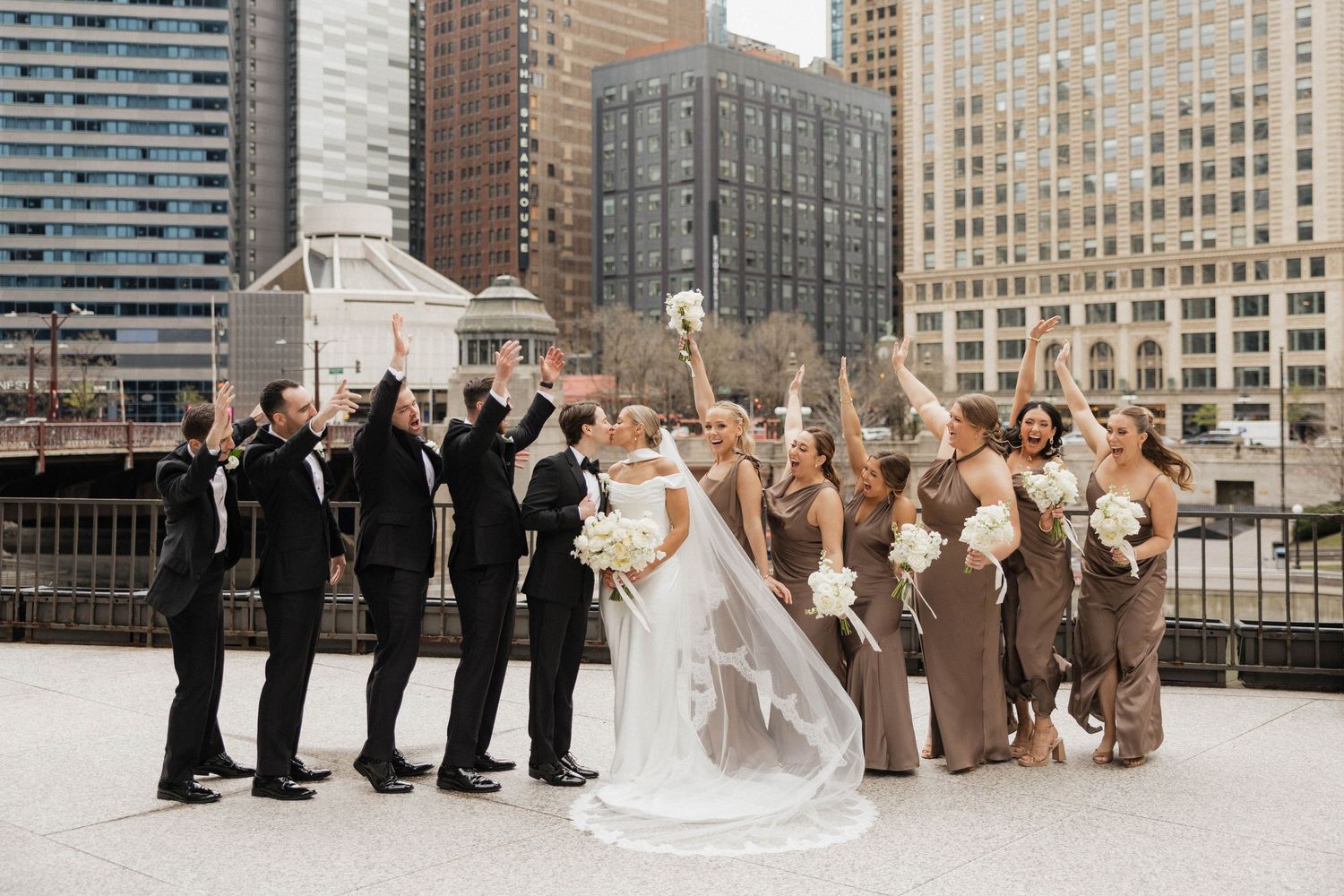 Wedding party celebrates on rooftop with Chicago skyline backdrop, bridesmaids in taupe dresses, groomsmen in black suits.
