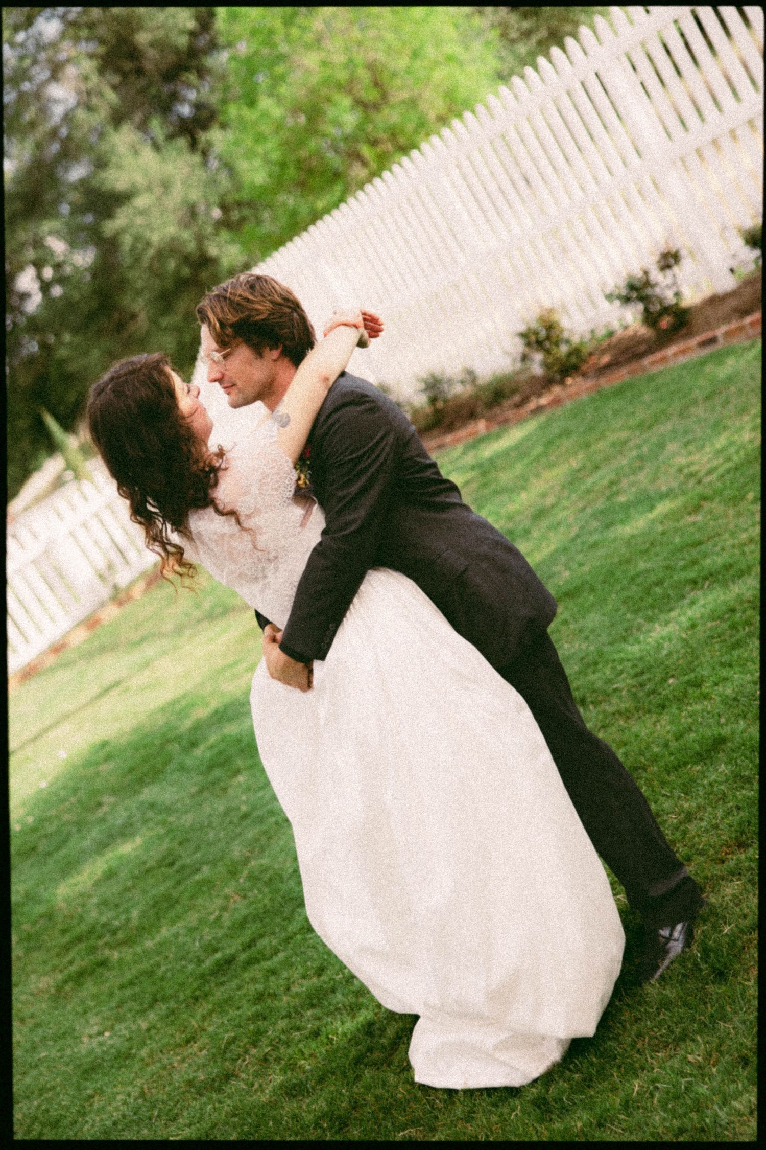 Bride and groom sharing romantic dip kiss on green lawn with white picket fence at outdoor wedding.