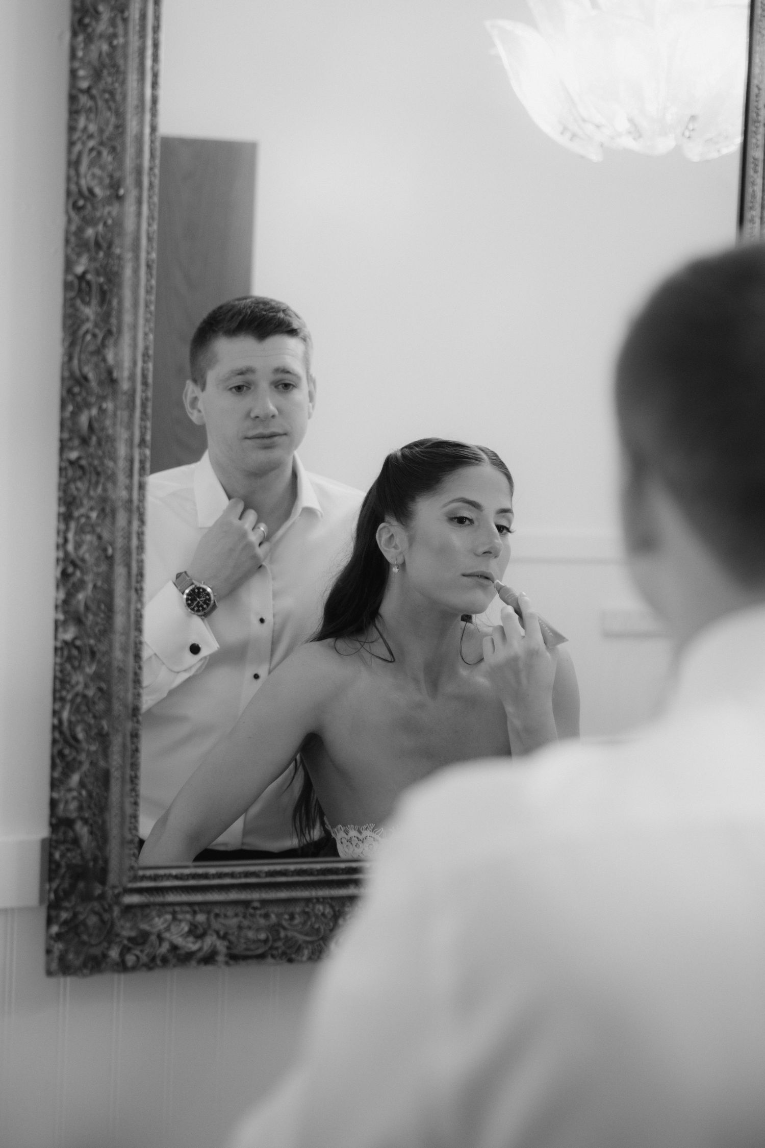 Black and white photo of a man helping a woman with her necklace in front of an ornate mirror.