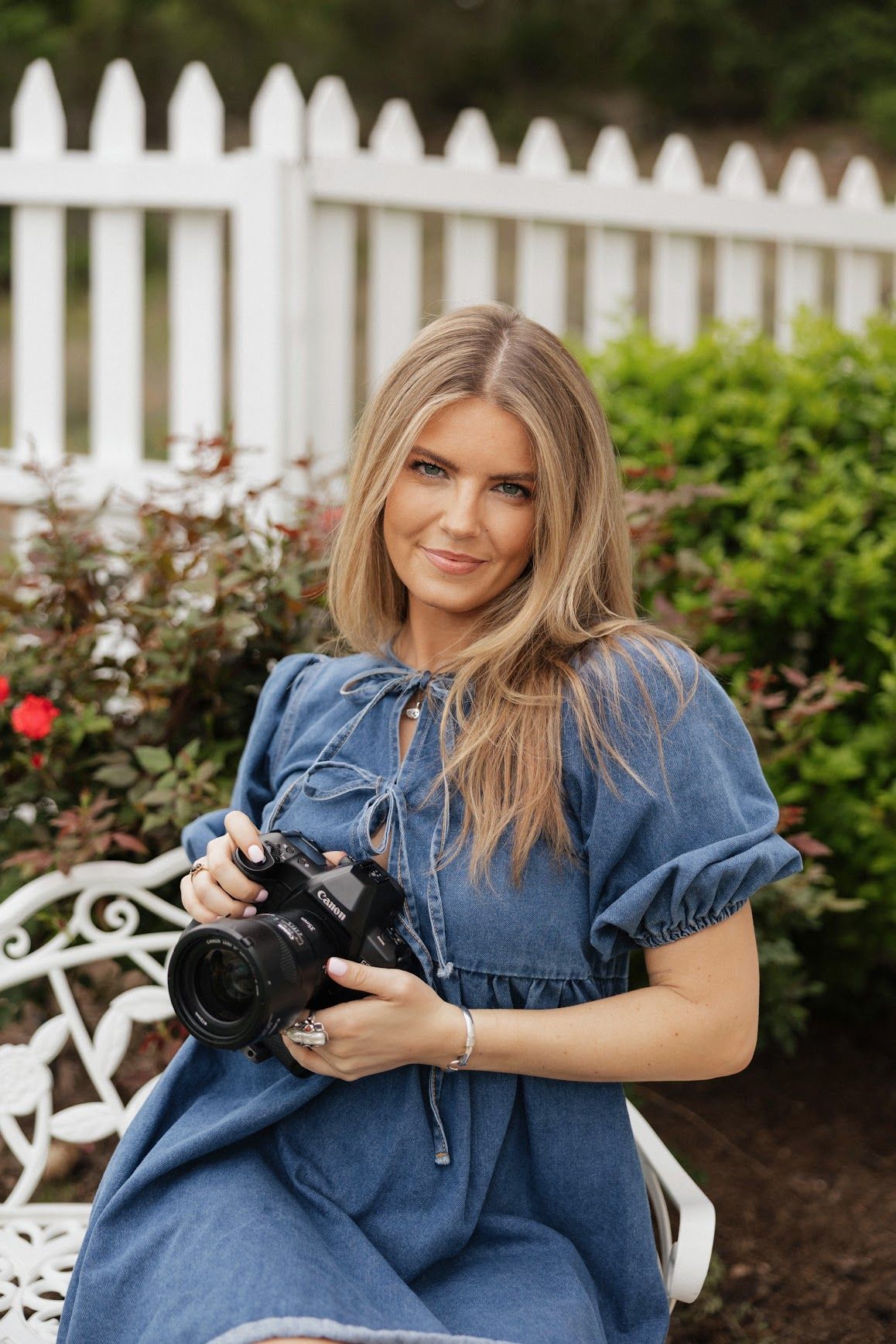 Blonde woman in blue denim dress holding a DSLR camera, seated outdoors near white picket fence and green shrubs.