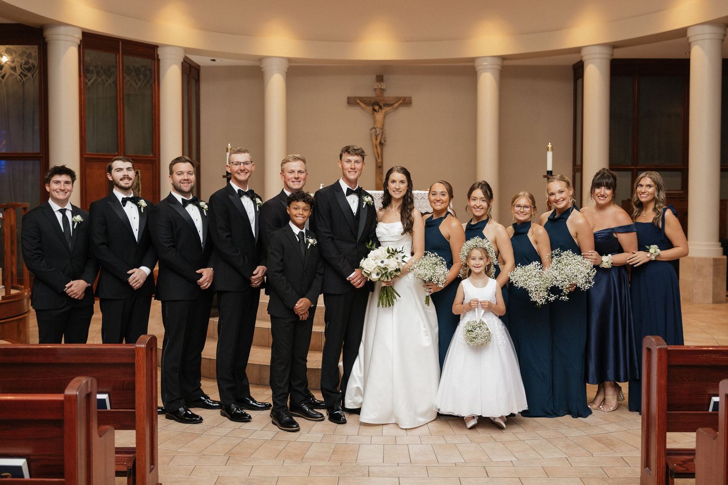 Wedding party poses in church with groomsmen in black tuxedos and bridesmaids in navy dresses flanking bride and groom.