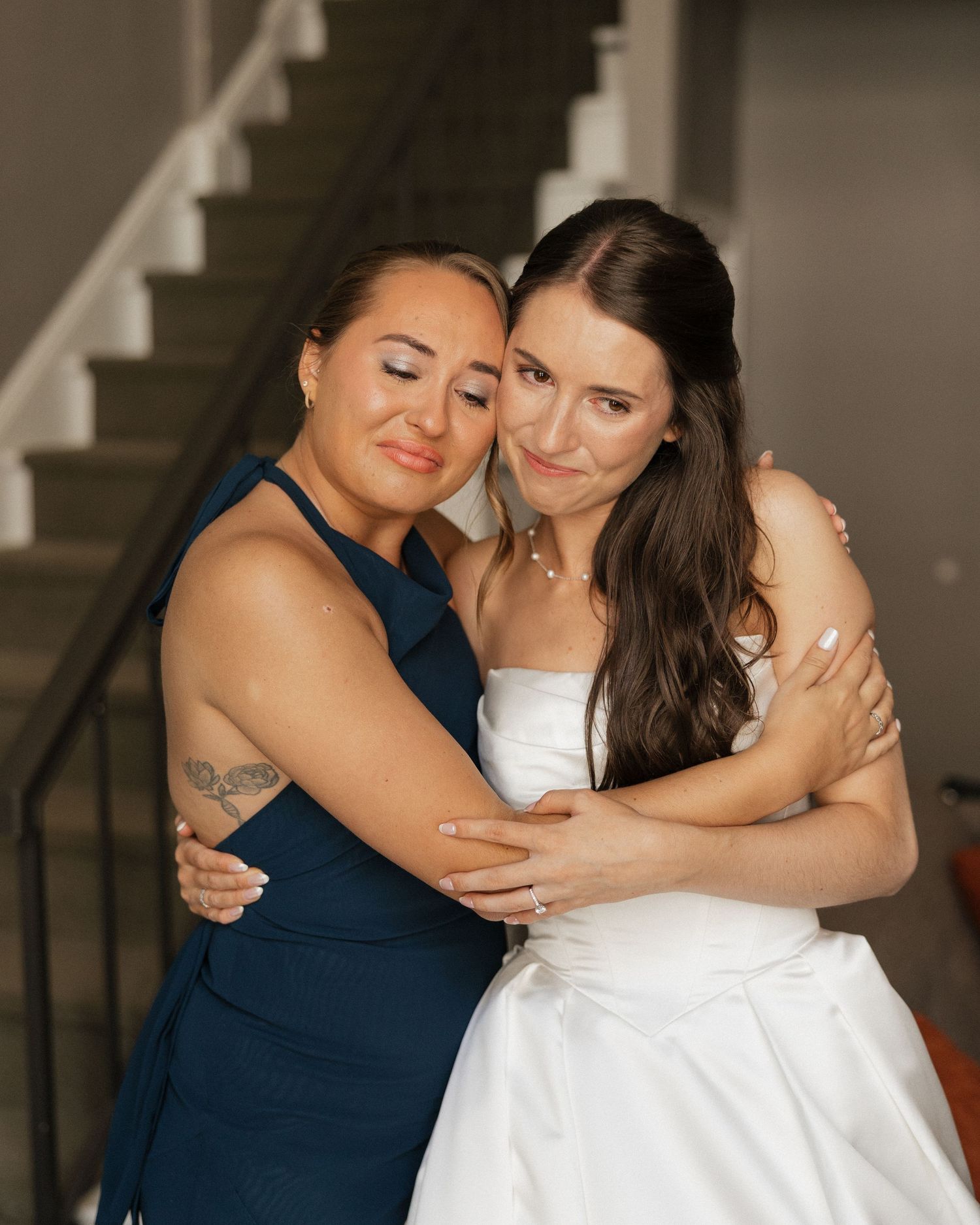 A bride in white gown and bridesmaid in navy dress share an emotional hug near a staircase.