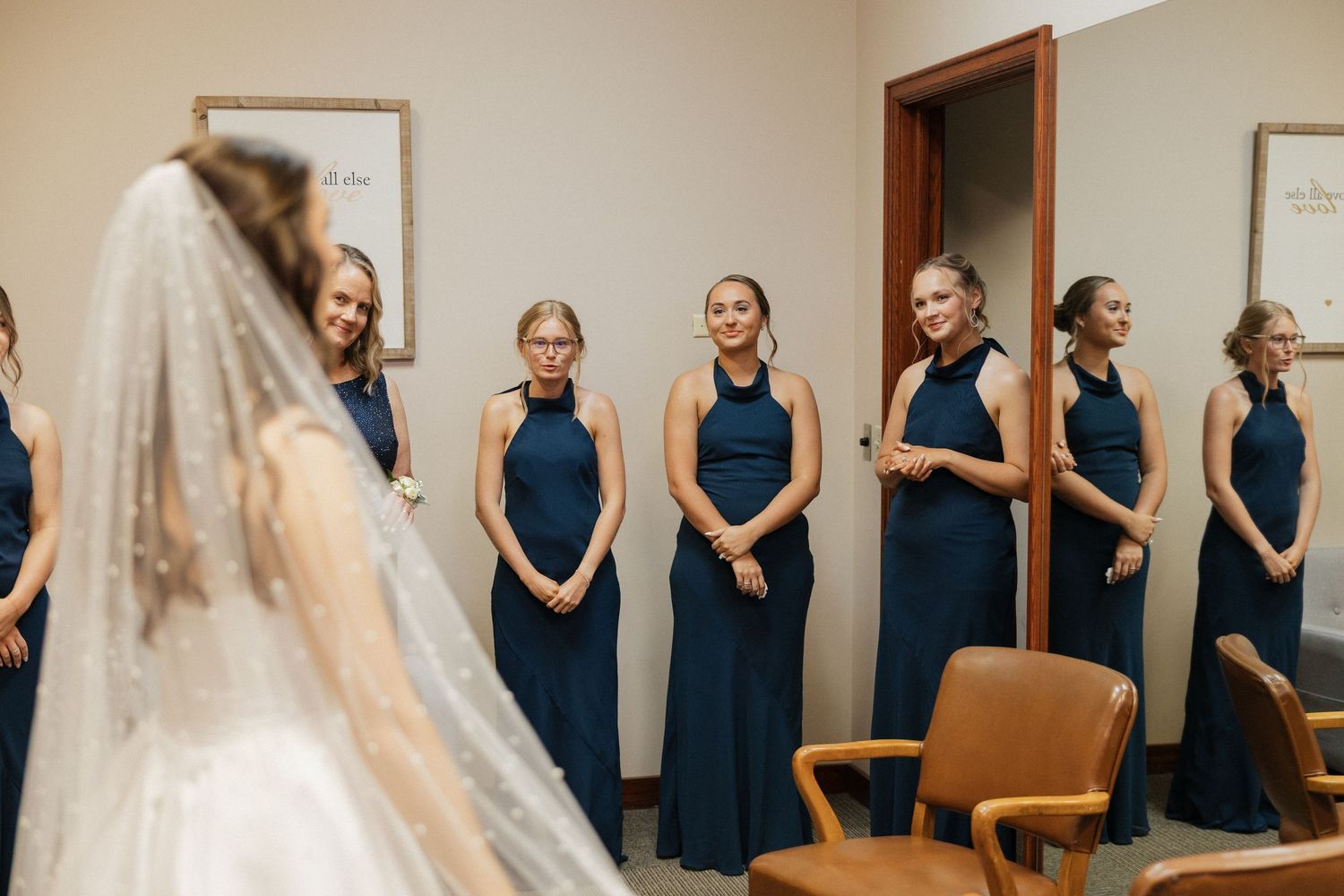 Bridesmaids in navy blue halter gowns smile at bride in white wedding dress and veil during bridal reveal.