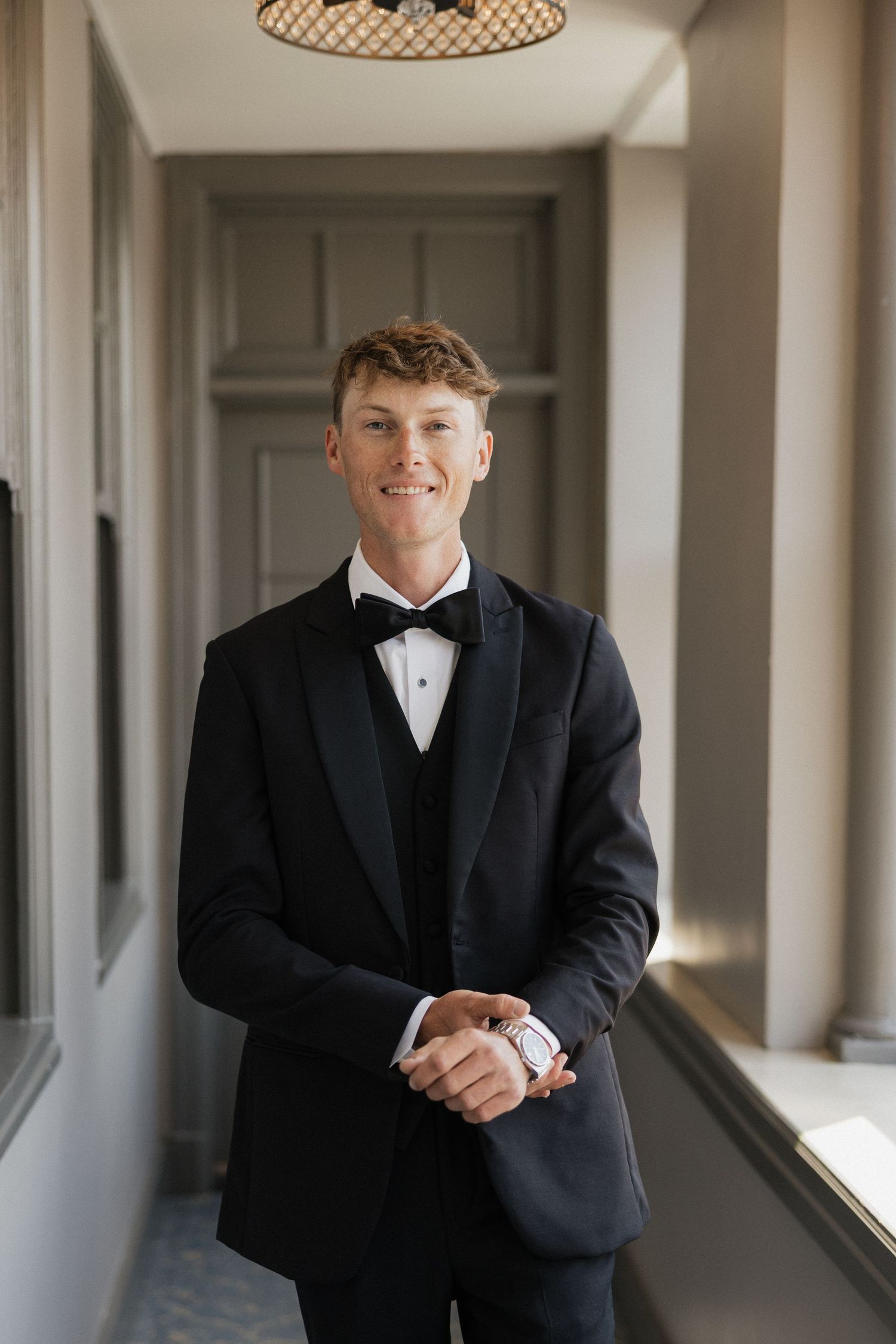 Young man in black tuxedo with bow tie smiling in elegant hallway, formal attire for wedding or prom.