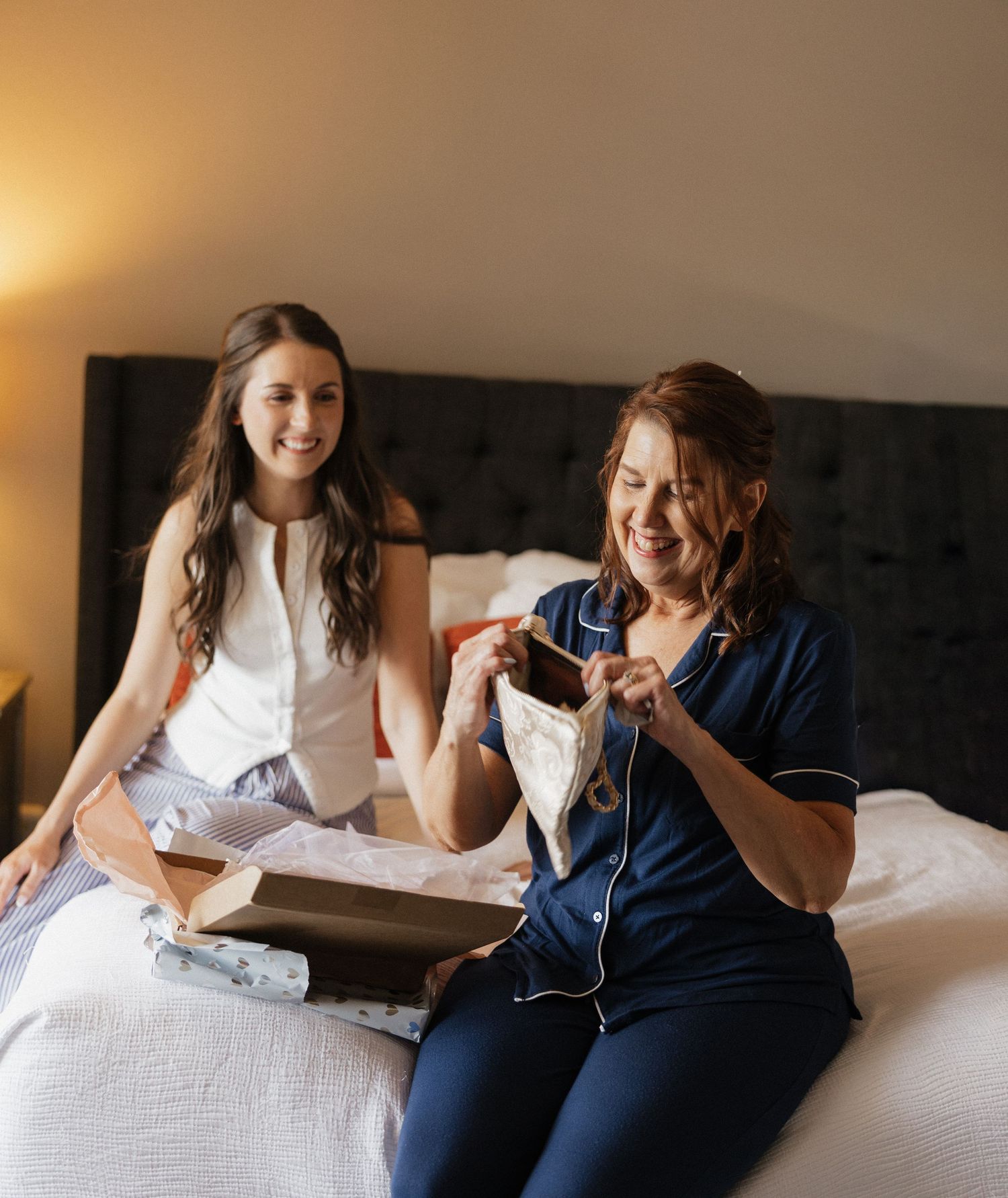 Two women sitting on a white bed, smiling while opening gifts, one wearing navy pajamas and the other a white sleeveless top.