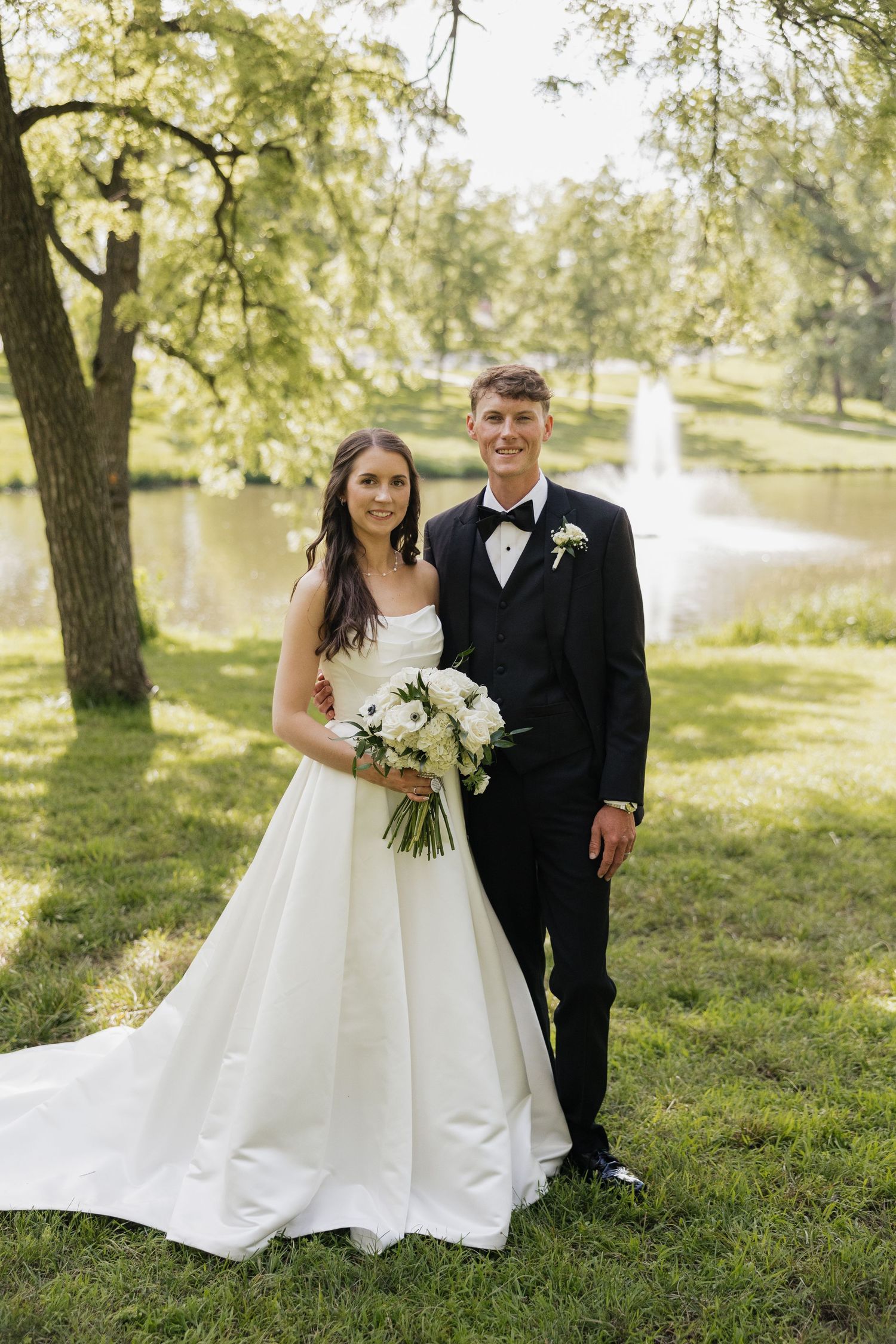 A bride in a white gown and groom in a black tuxedo pose together outdoors near a pond in a lush green park.