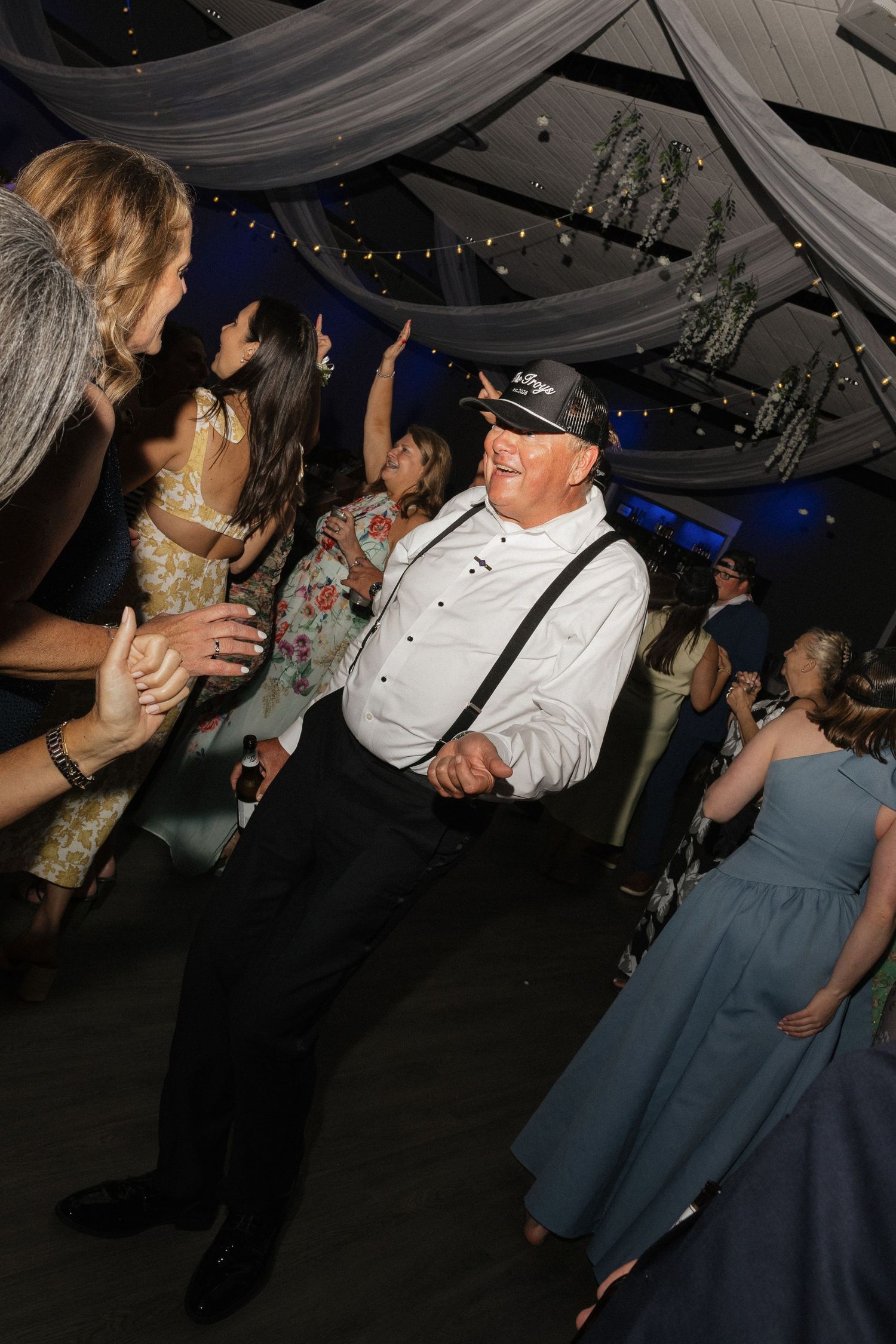 Elderly man in white shirt, suspenders, and cap dancing joyfully at an elegant wedding reception.