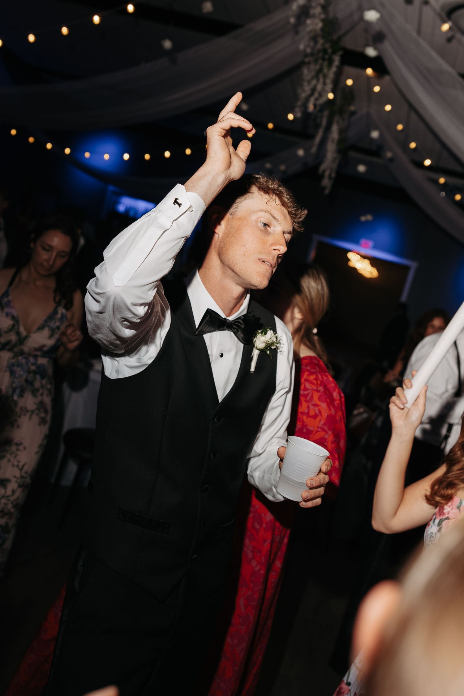 A groom in a black tuxedo vest and bow tie dances energetically at a wedding reception, pointing up while holding a cup.