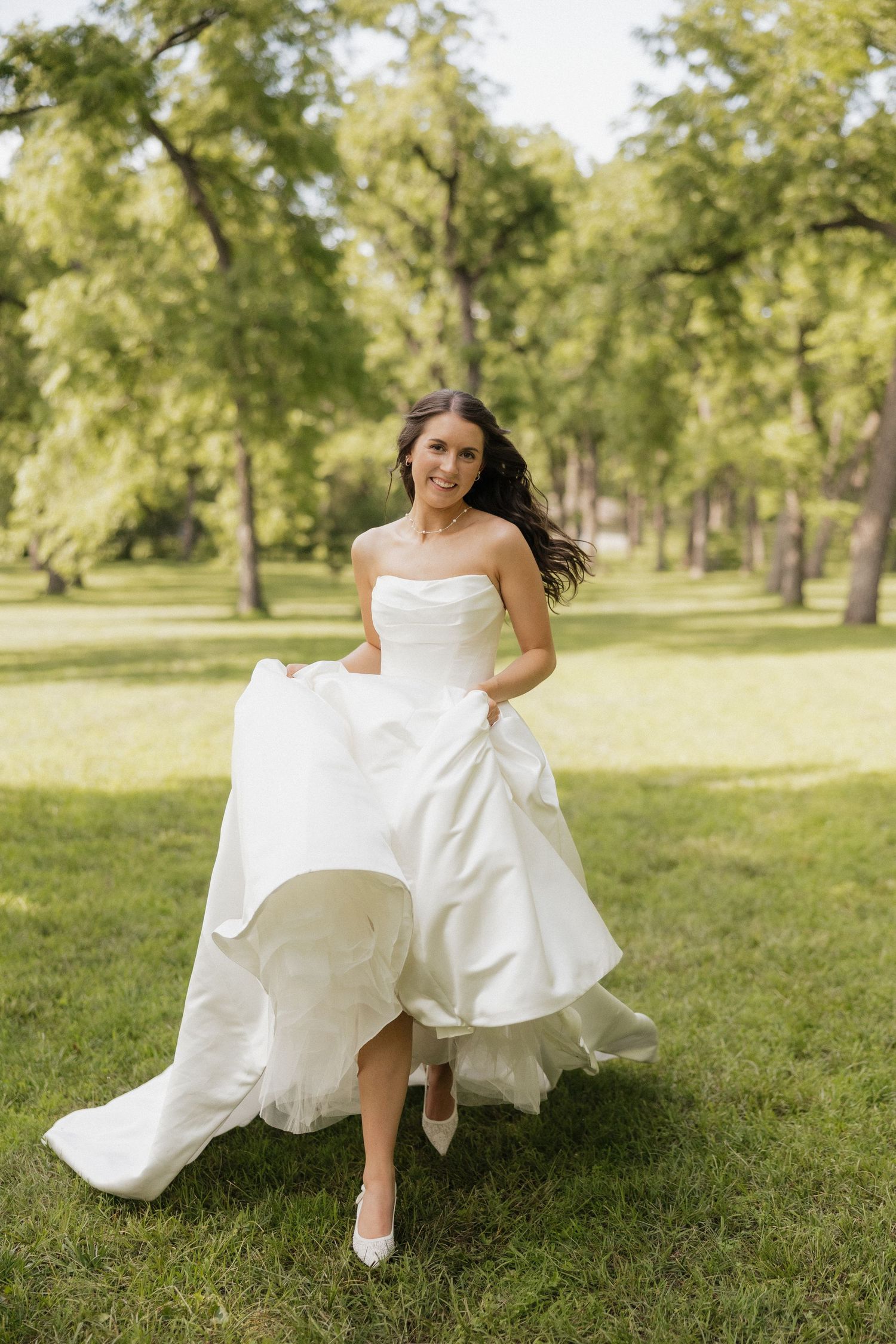 Smiling bride in white strapless ballgown running through green park, dark hair flowing, wearing white heels on sunny day.