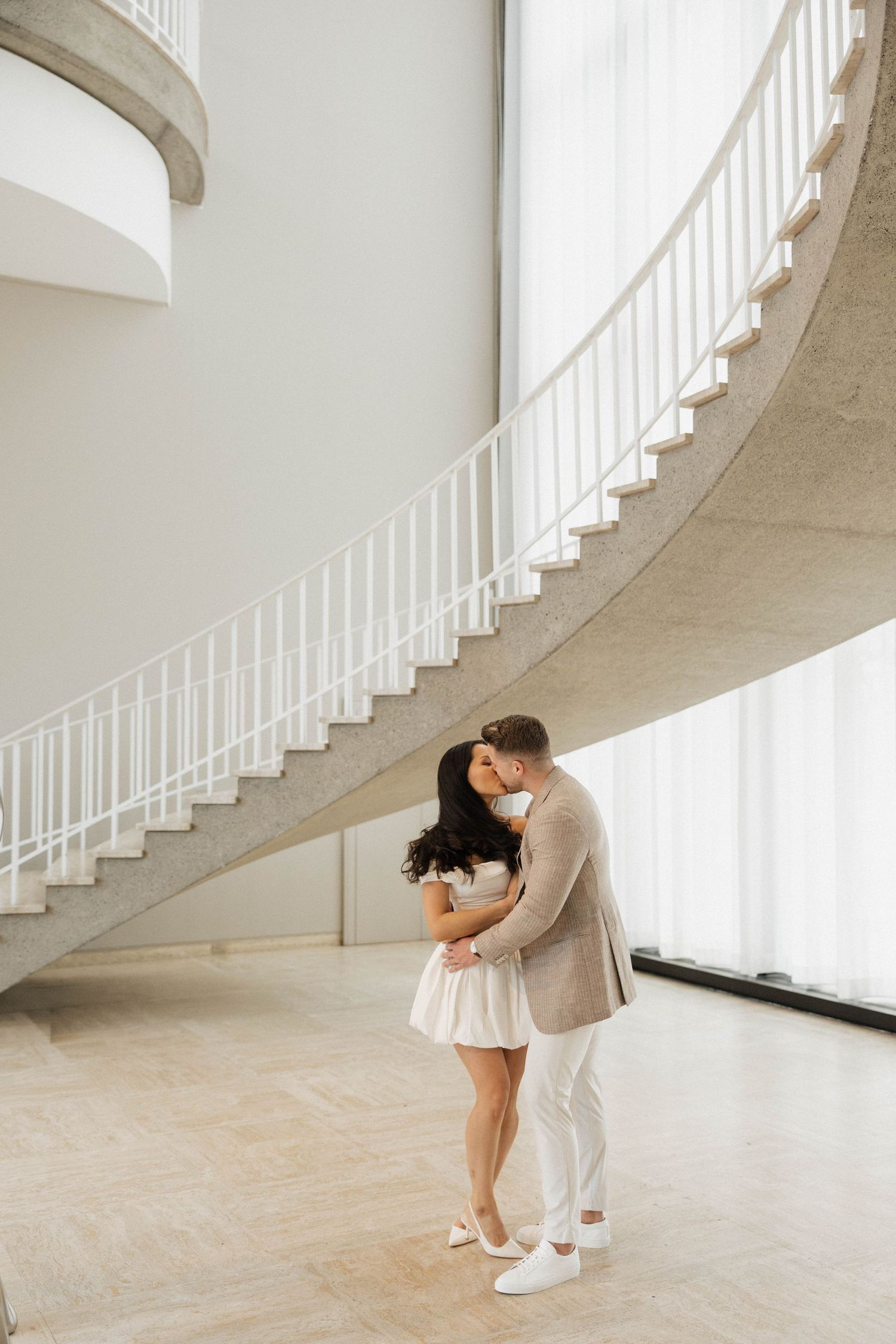 Couple embracing tenderly beneath an elegant curved spiral staircase in a bright modern building interior.