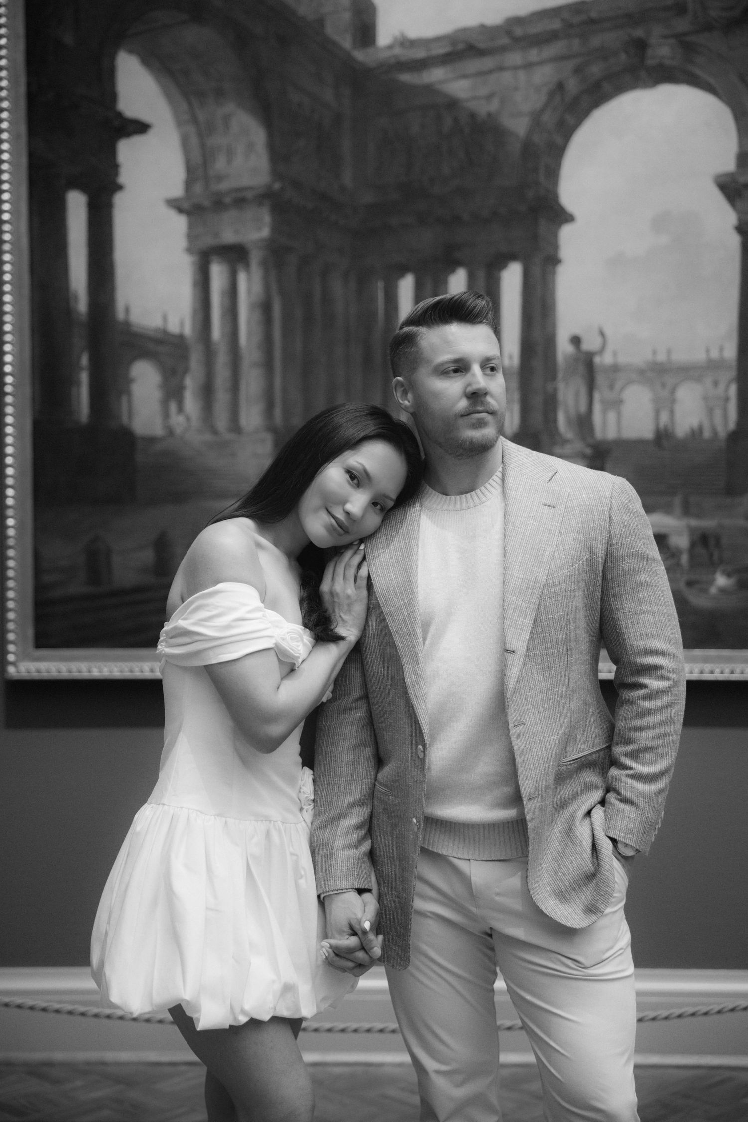 Black and white photo of a couple posing in an art museum, woman in white dress leaning on mans shoulder.