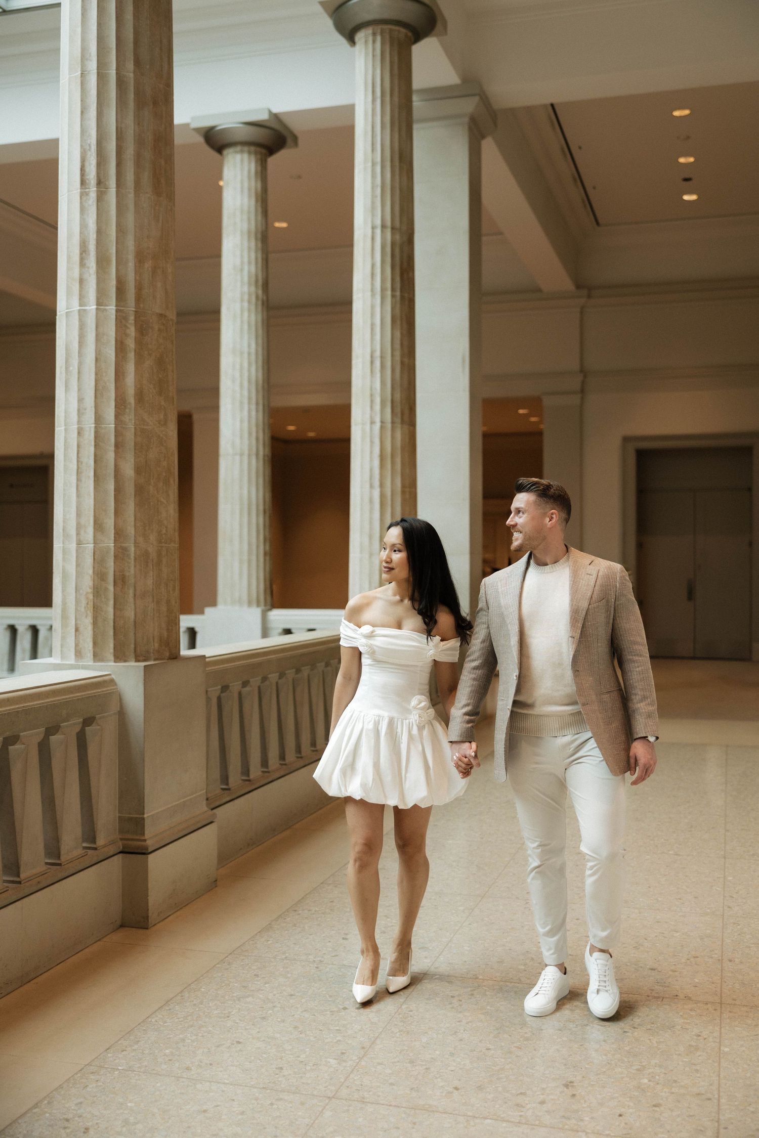 Romantic couple holding hands and smiling at each other while walking through an elegant columned museum gallery.