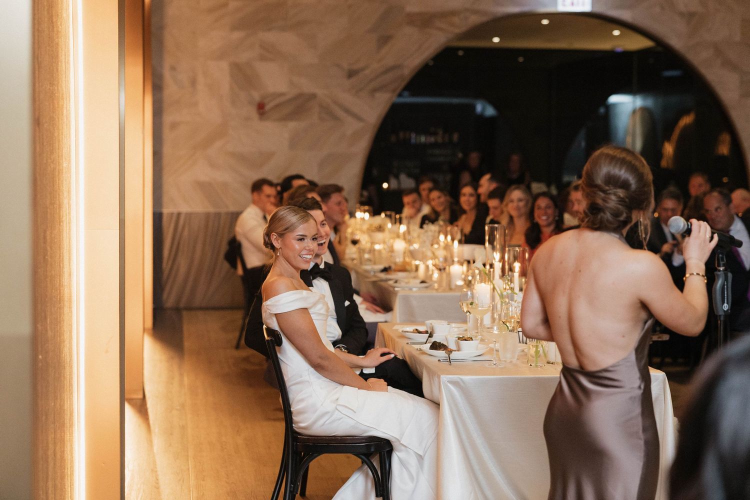 Bride smiling at candlelit wedding reception table while bridesmaid in taupe dress stands nearby in elegant venue.