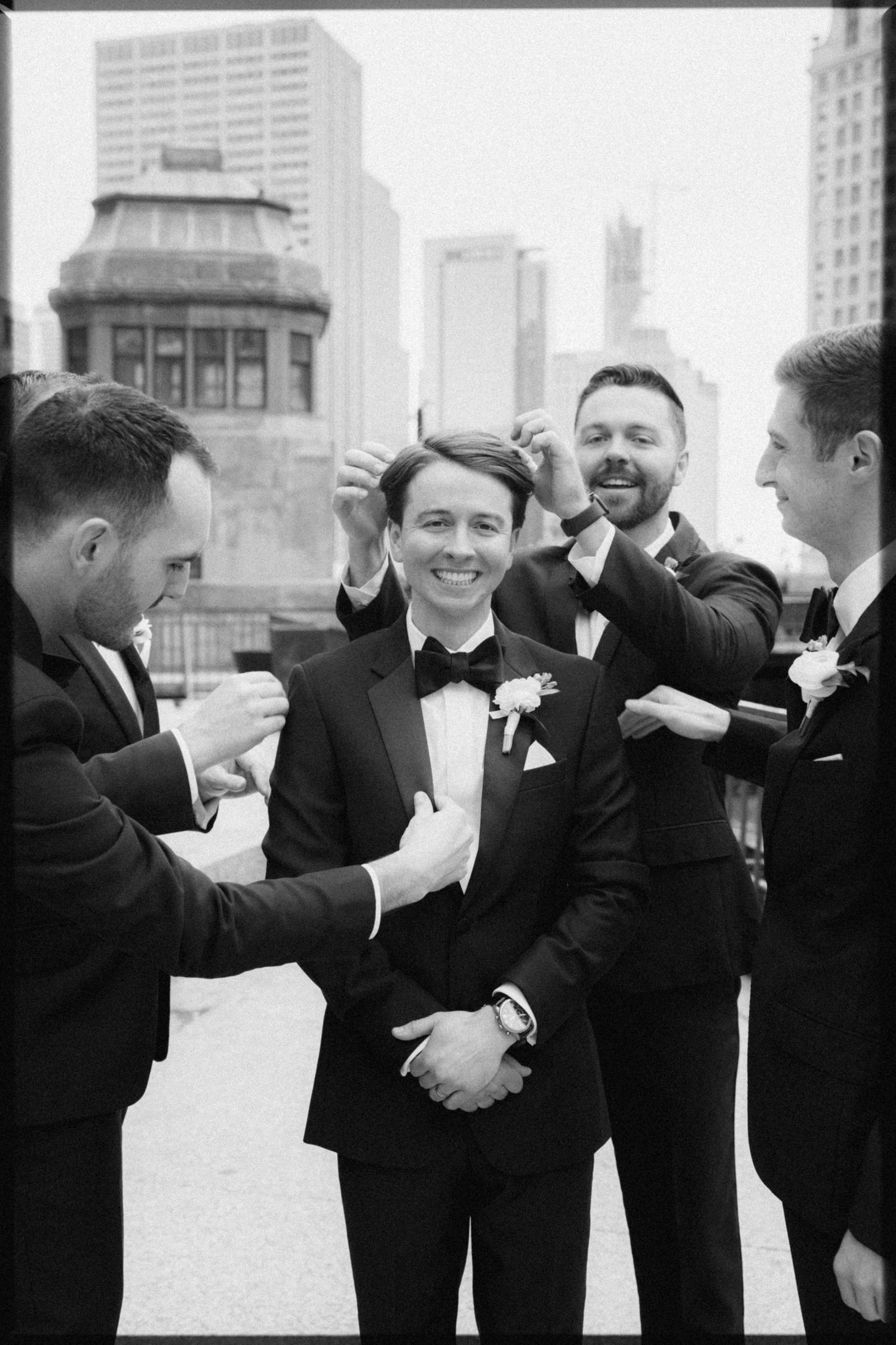 Groomsmen adjusting groom's suit and hair outdoors with city skyline in background on wedding day.