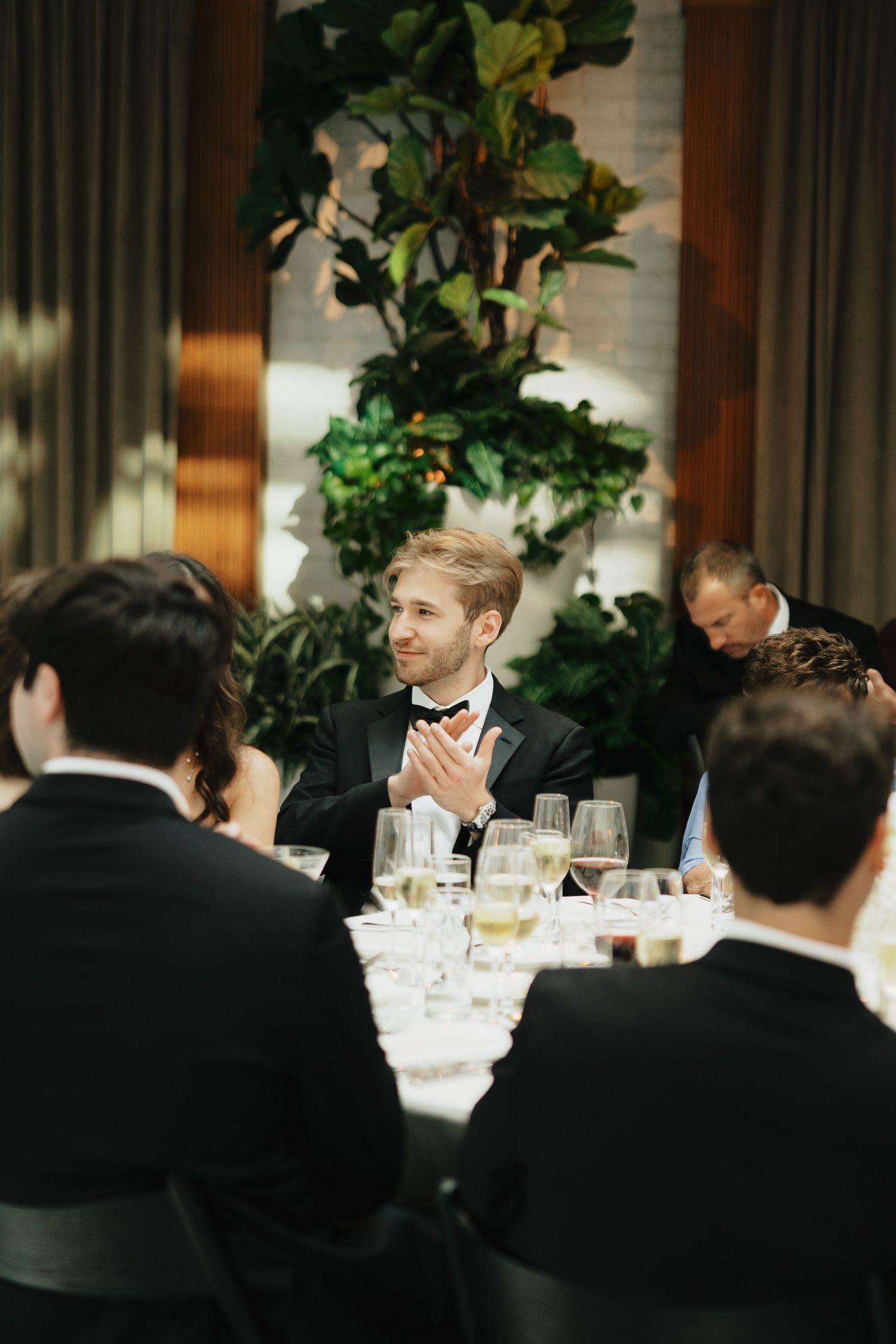 Groom in black tuxedo clapping at elegant wedding reception dinner table with guests and lush green plants.