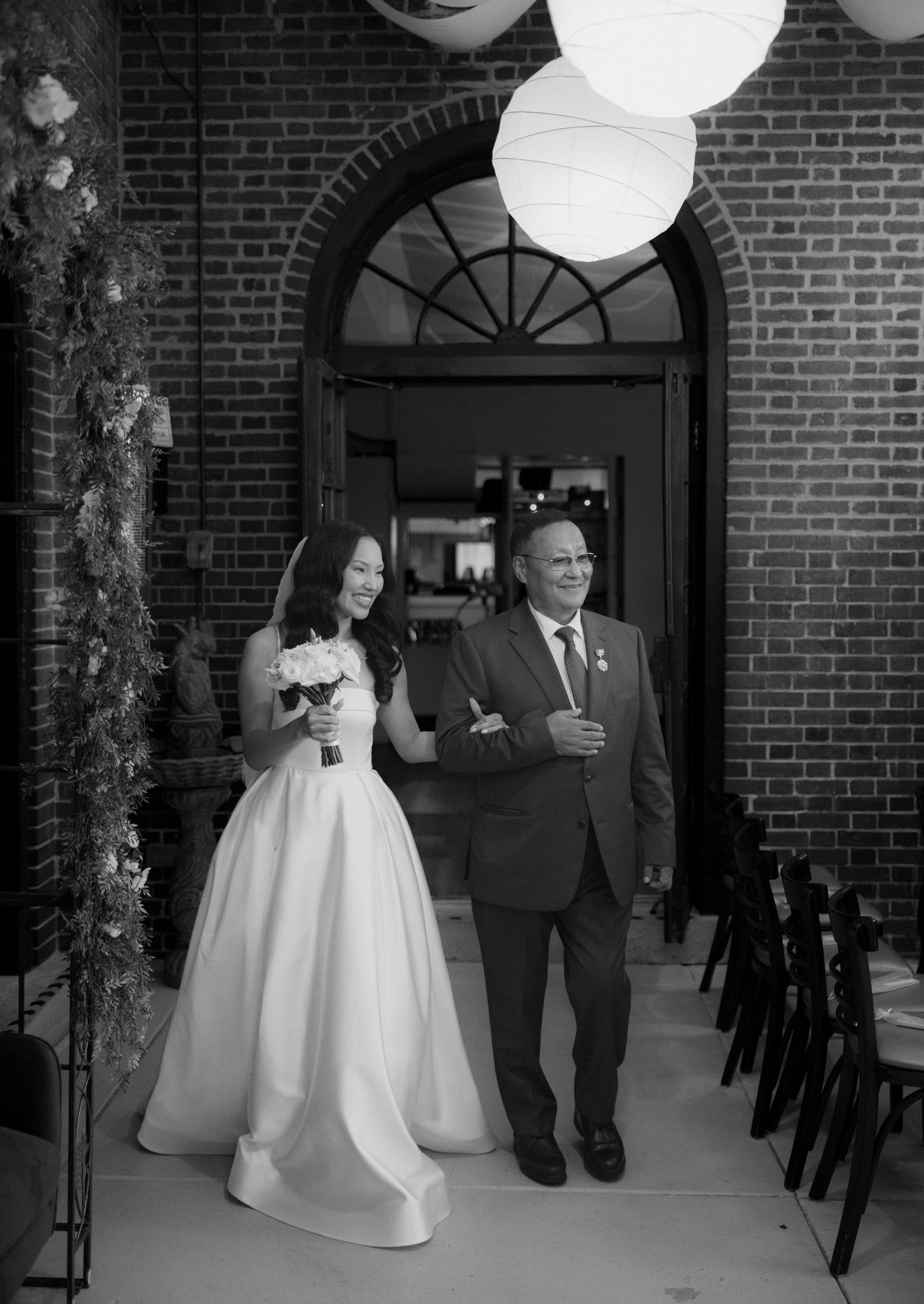 Black and white photo of a bride in a gown holding a bouquet, arm in arm with an older man in a suit at a brick archway entrance.