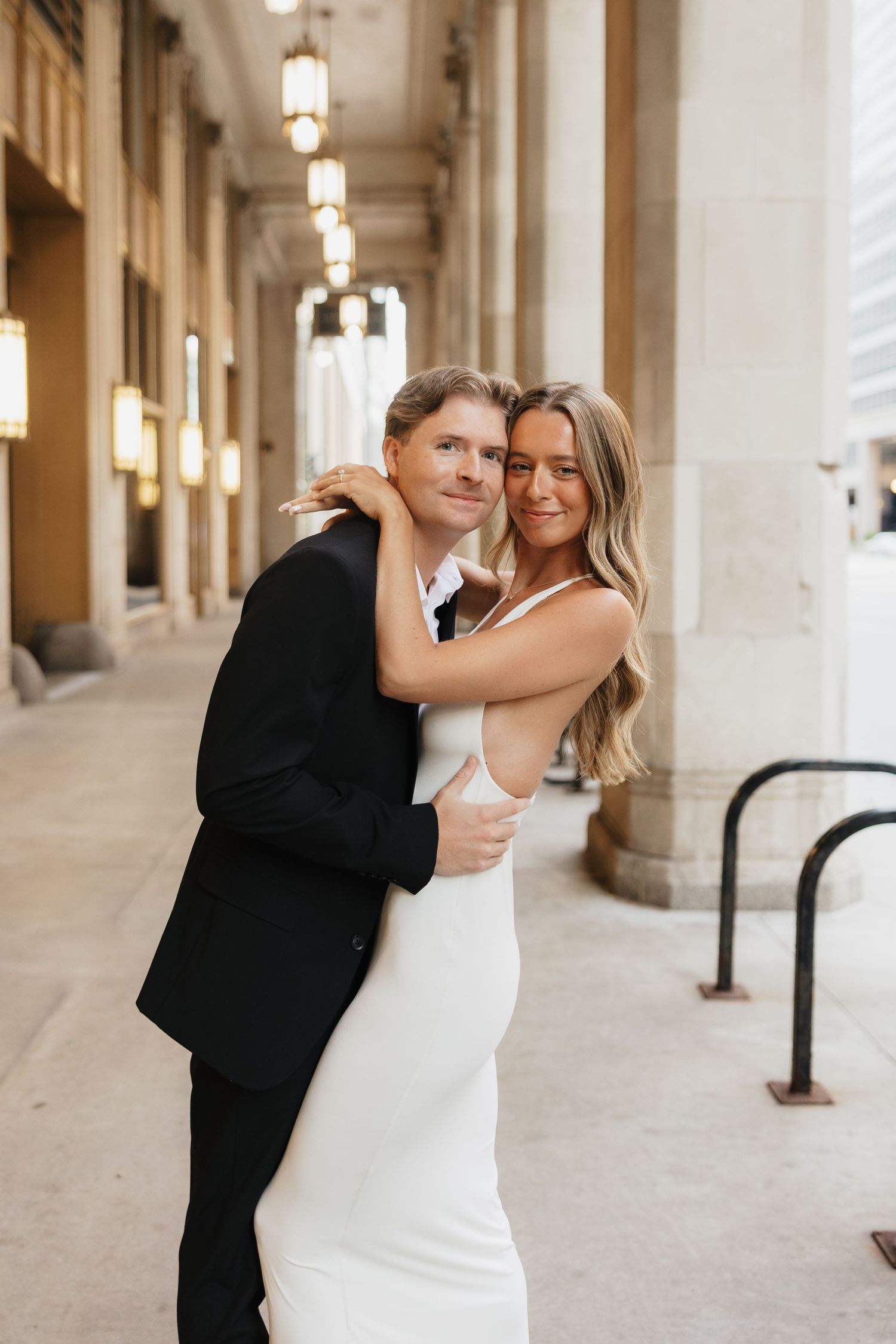 A couple embraces in an elegant white dress and black suit beneath tall stone columns and hanging lanterns in a city archway.