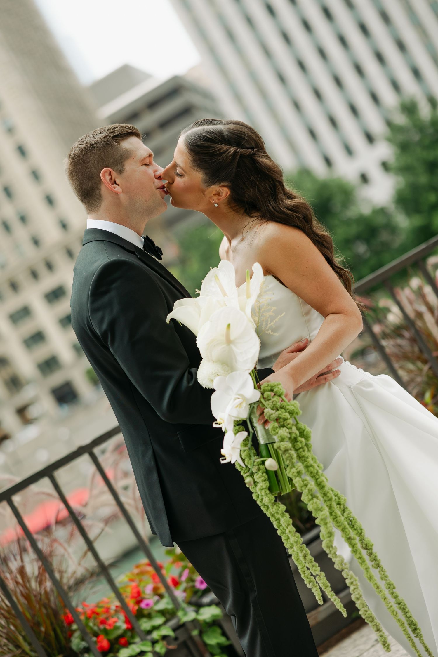 Bride in a white lace gown holding a cascading white calla lily and green bouquet kisses her groom in a black tuxedo on an urban balcony.