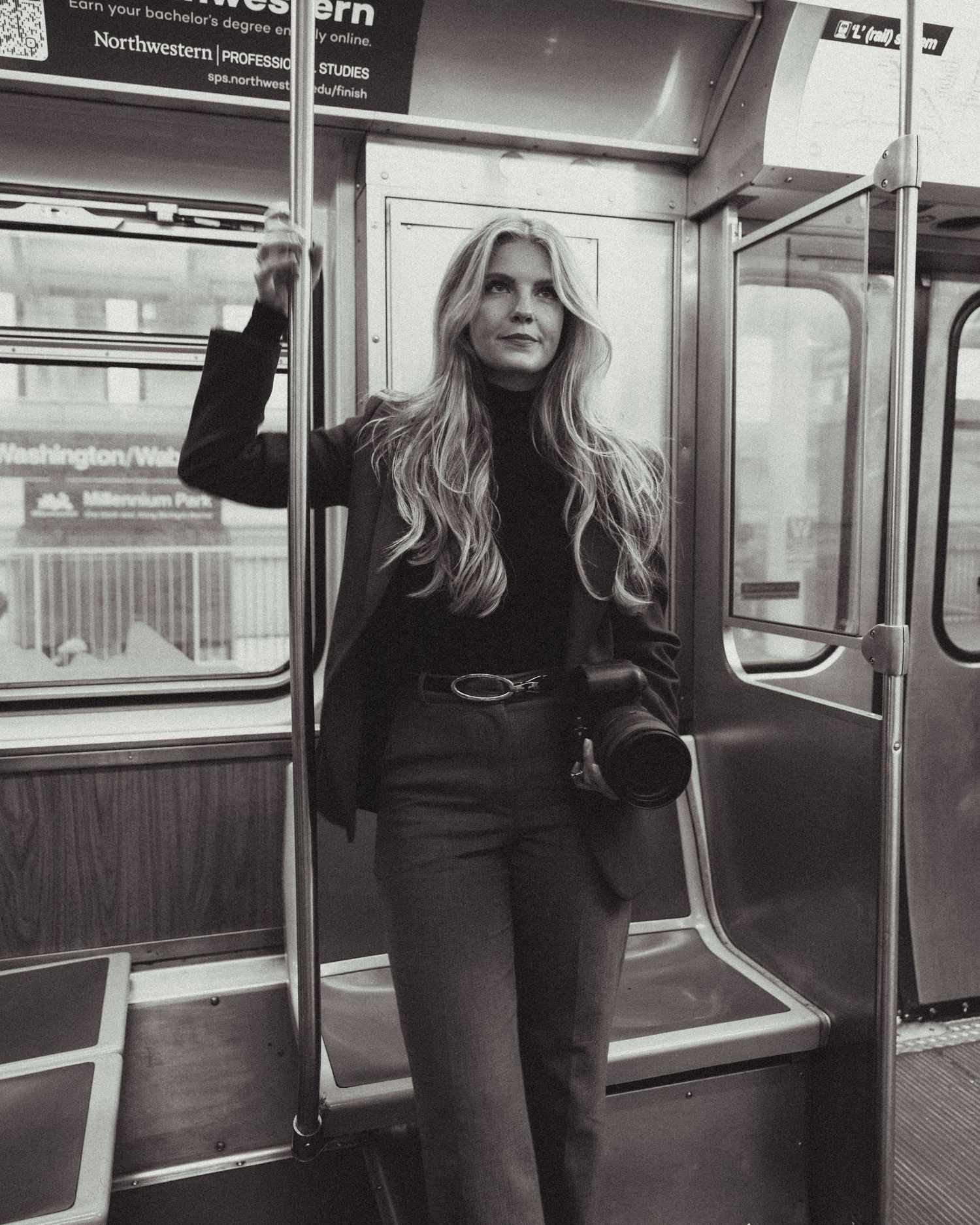 Black and white photo of a blonde woman in dark outfit holding camera on subway train at Washington station.