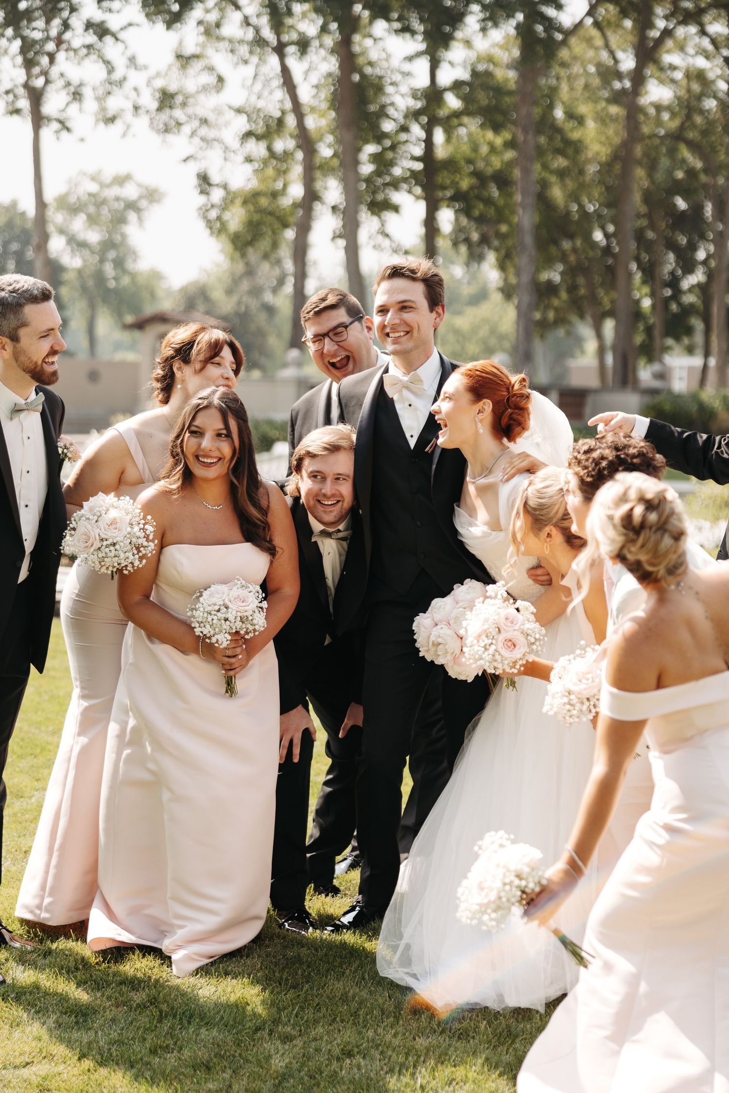 Wedding party laughing outdoors, bridesmaids in white gowns holding bouquets with groomsmen in black suits on green lawn.