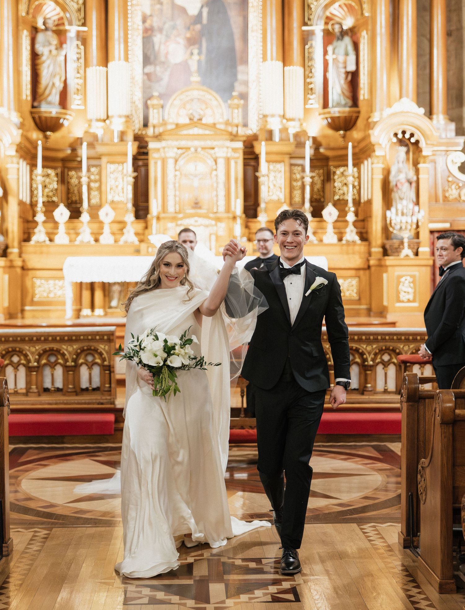 Newlyweds celebrate joyfully while walking down the aisle of an ornate cathedral with golden altar and decorative details.