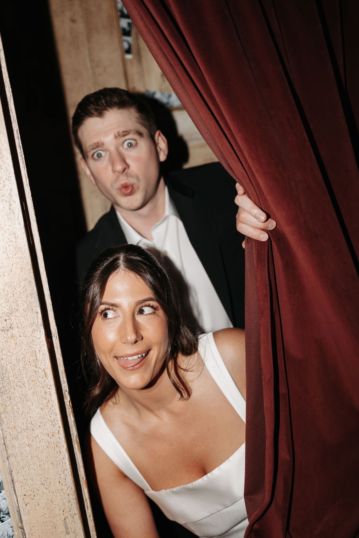 A playful couple peeks through a red curtain with exaggerated expressions during what appears to be their wedding day.