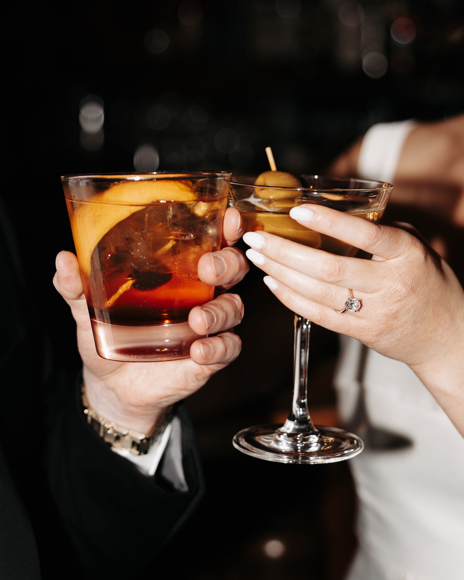 Multiple hands holding cocktail glasses with orange garnishes in a celebratory toast against a dark background.