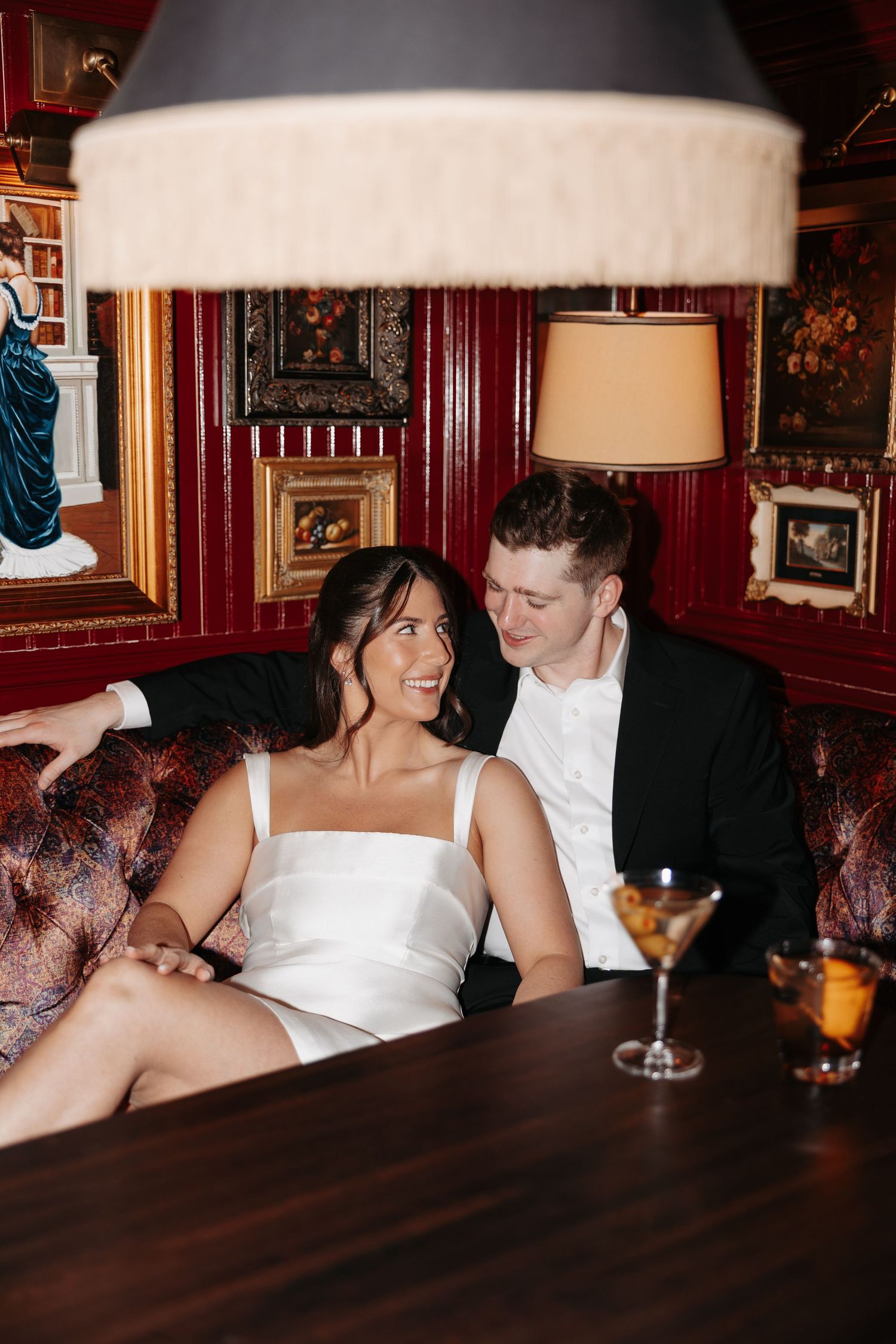 A couple shares a joyful moment in a vintage-style bar with red walls and ornate frames, martini glass on the table.