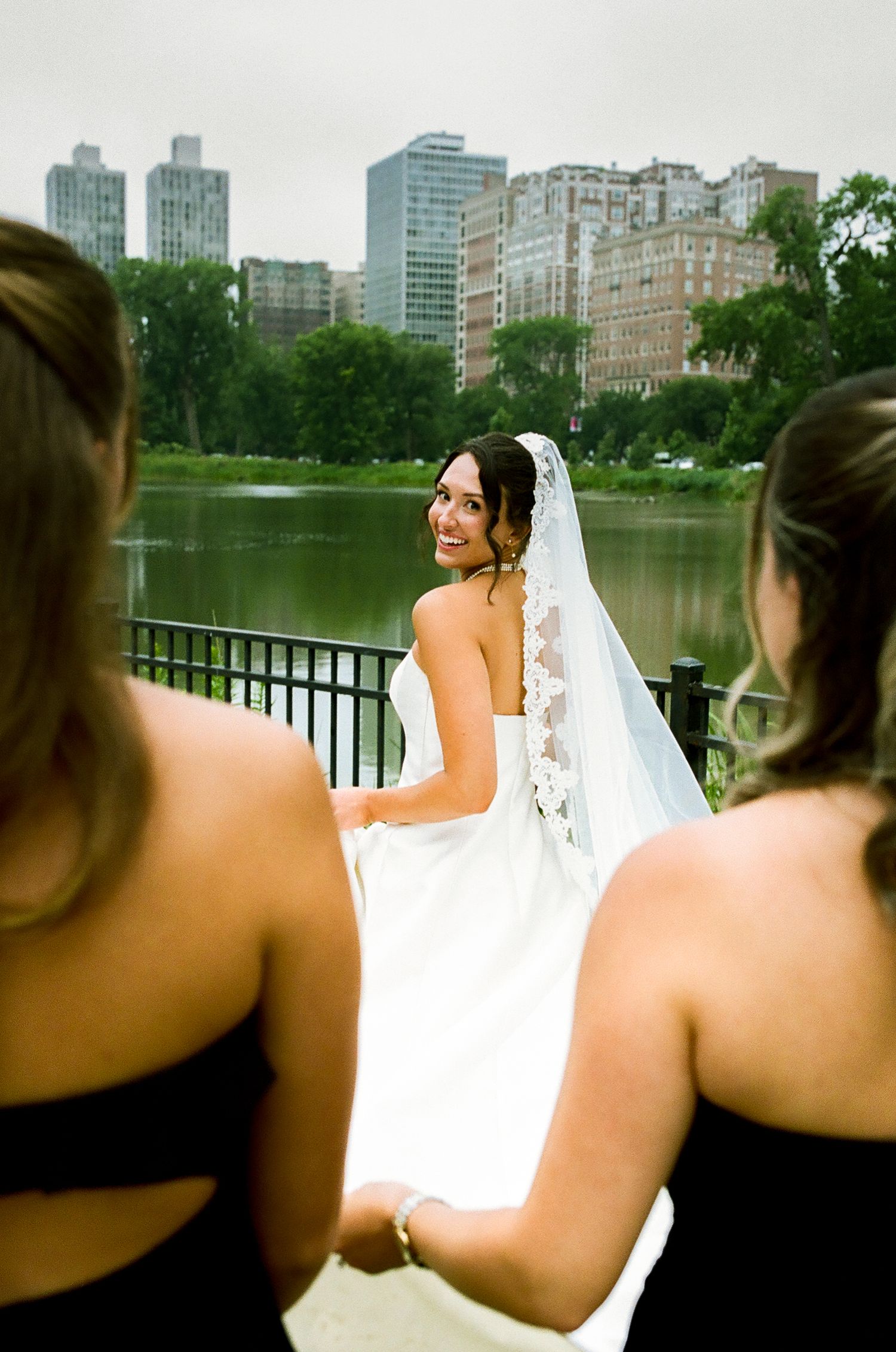 Bride in white gown and veil smiles at bridesmaids in black dresses near a lake with NYC skyline backdrop.