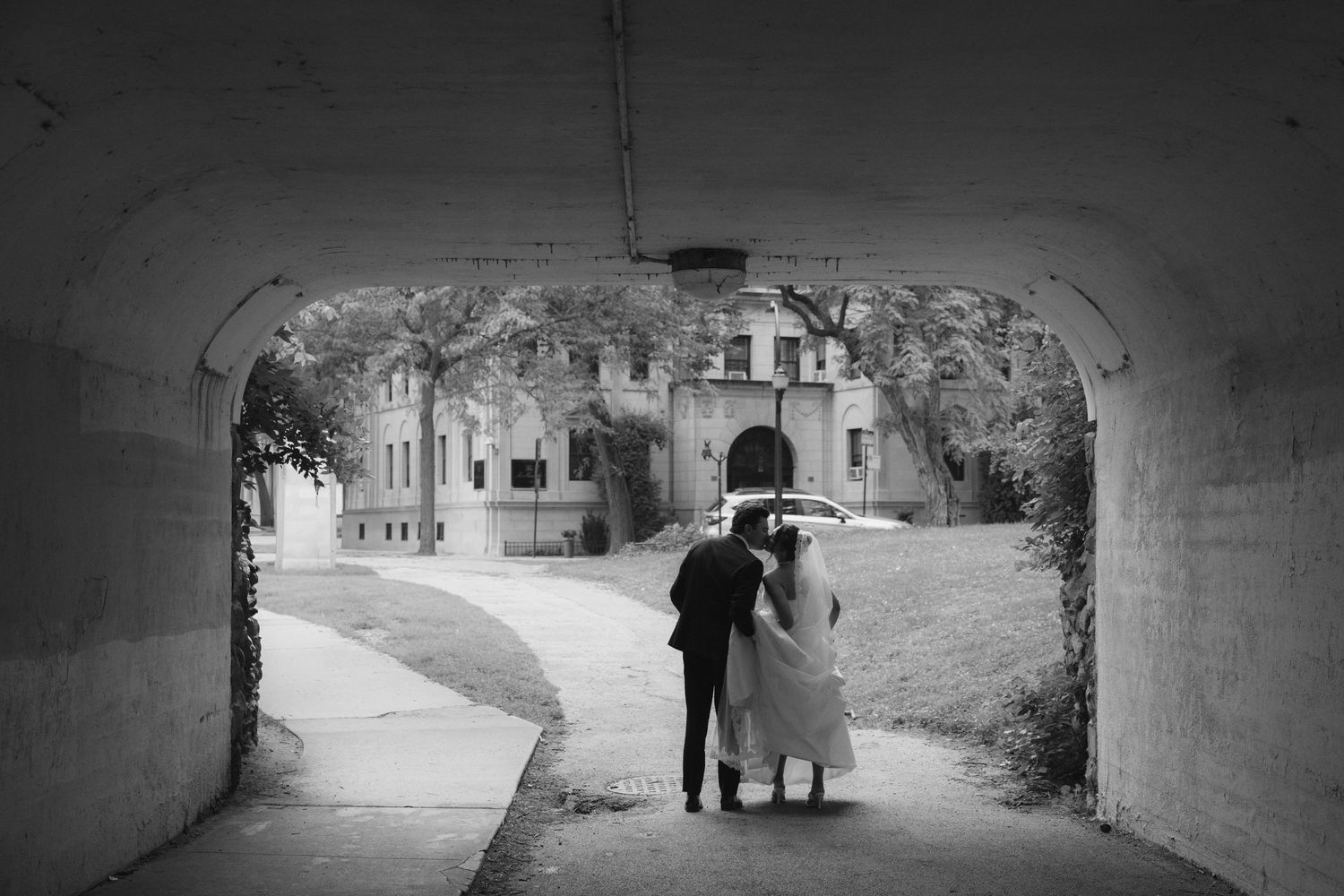 Black and white photo of a bride and groom walking through a tunnel arch toward a historic stone building.