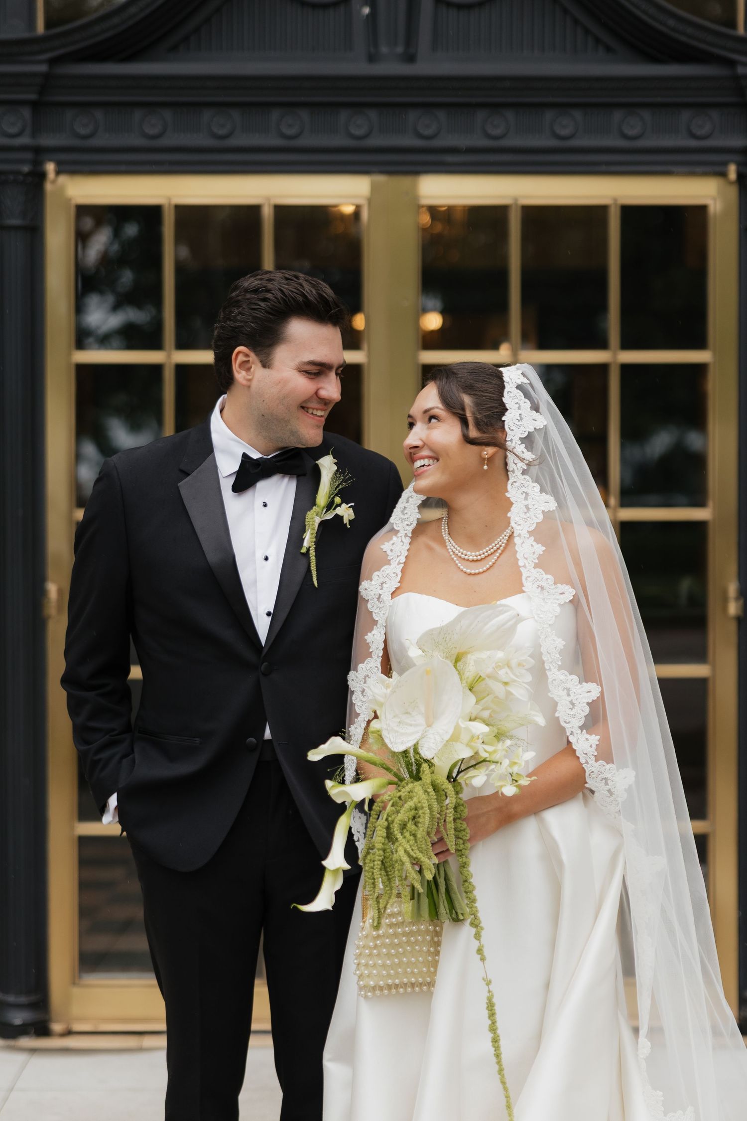 A smiling bride in a lace veil holds white calla lilies beside a tuxedo-clad groom outside elegant gold doors.