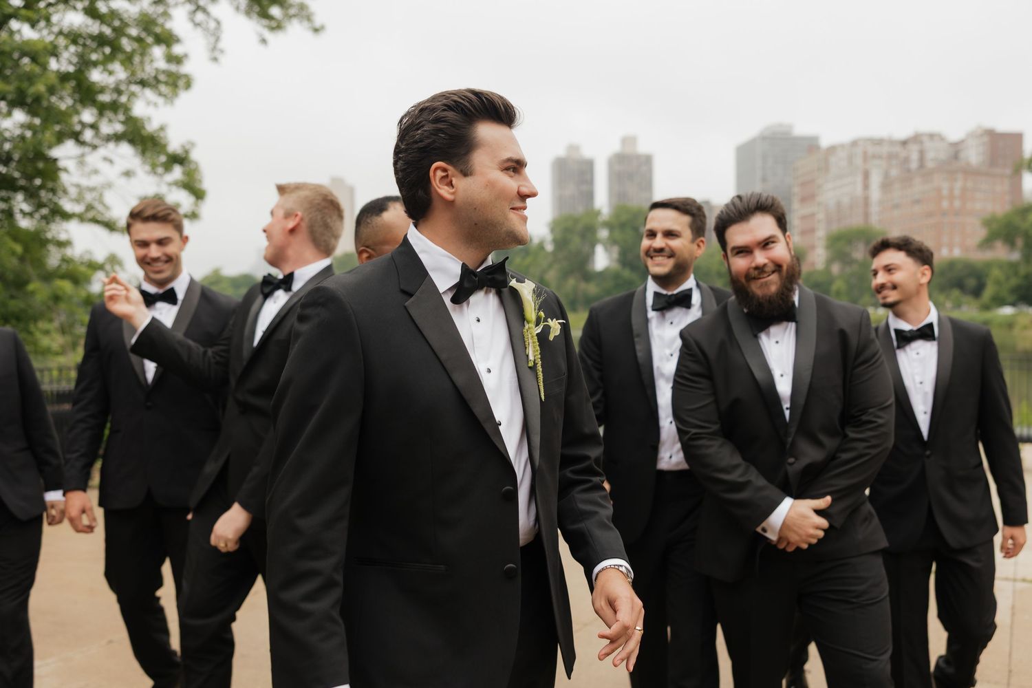 Groom in black tuxedo with boutonniere smiles with groomsmen in matching suits outdoors at wedding.