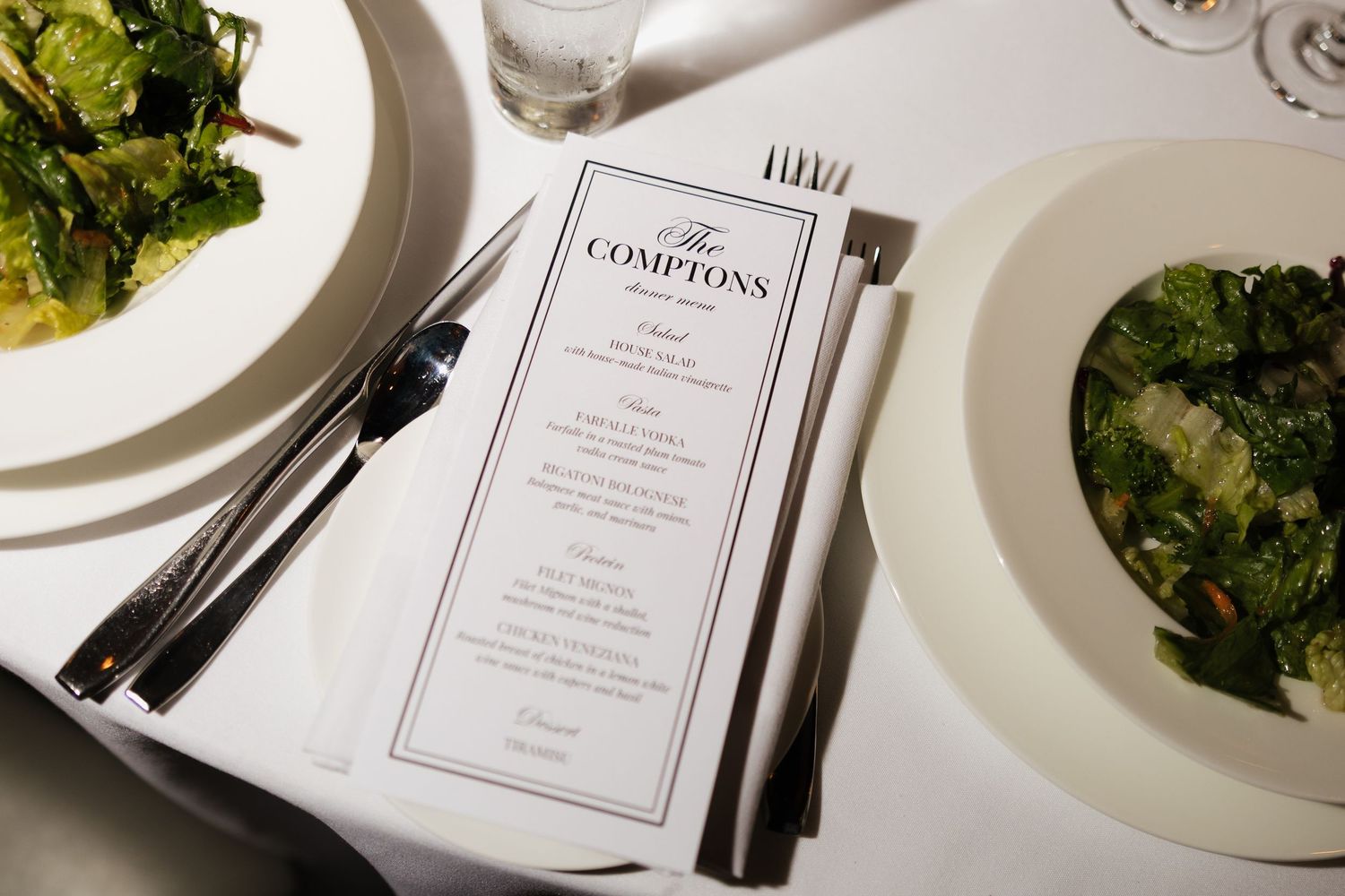 Elegant table setting at The Comptons restaurant with a menu card, silverware, and fresh salad plates on white linen.