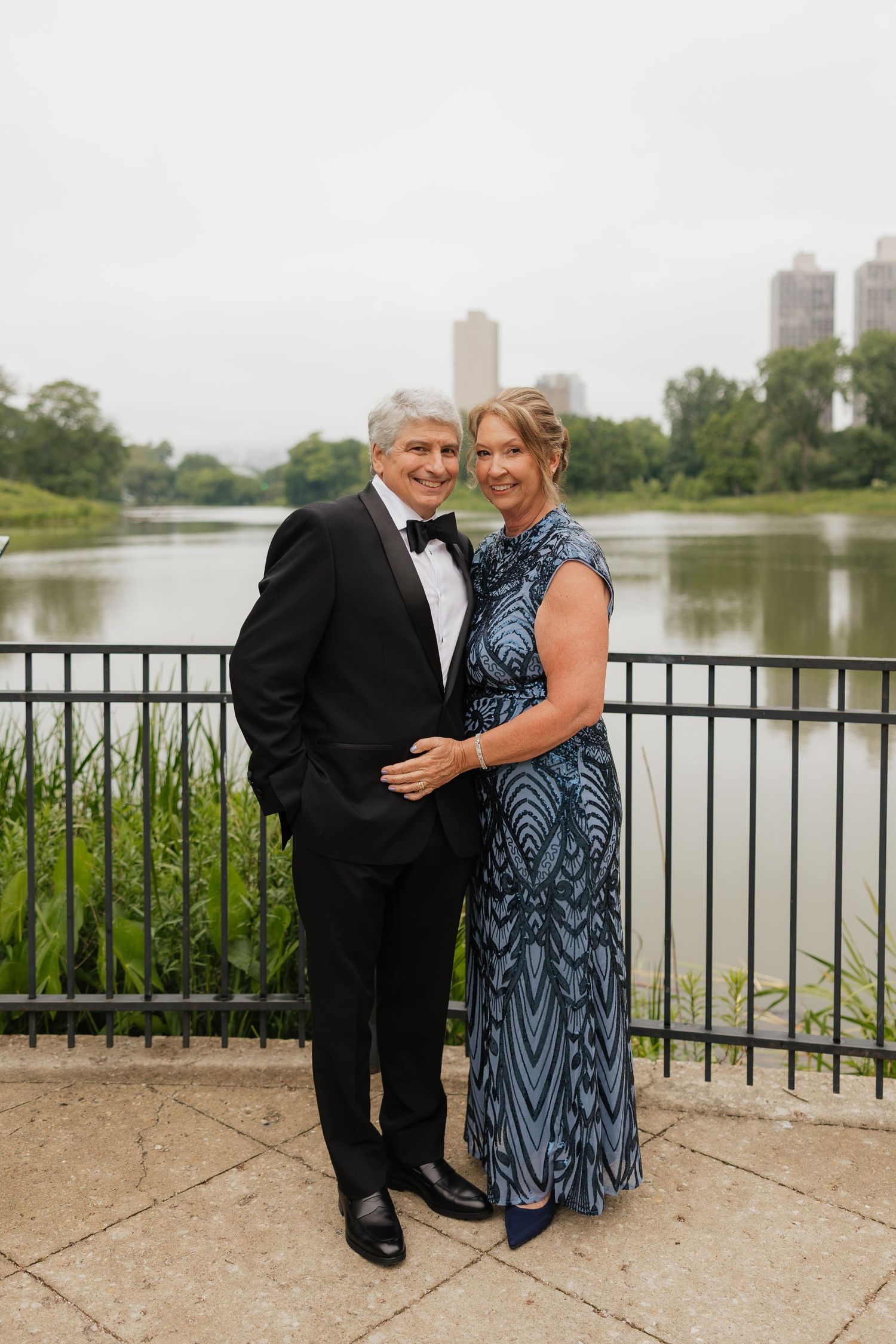 Elegant couple in formal attire posing by a lakeside railing with city skyline in background.