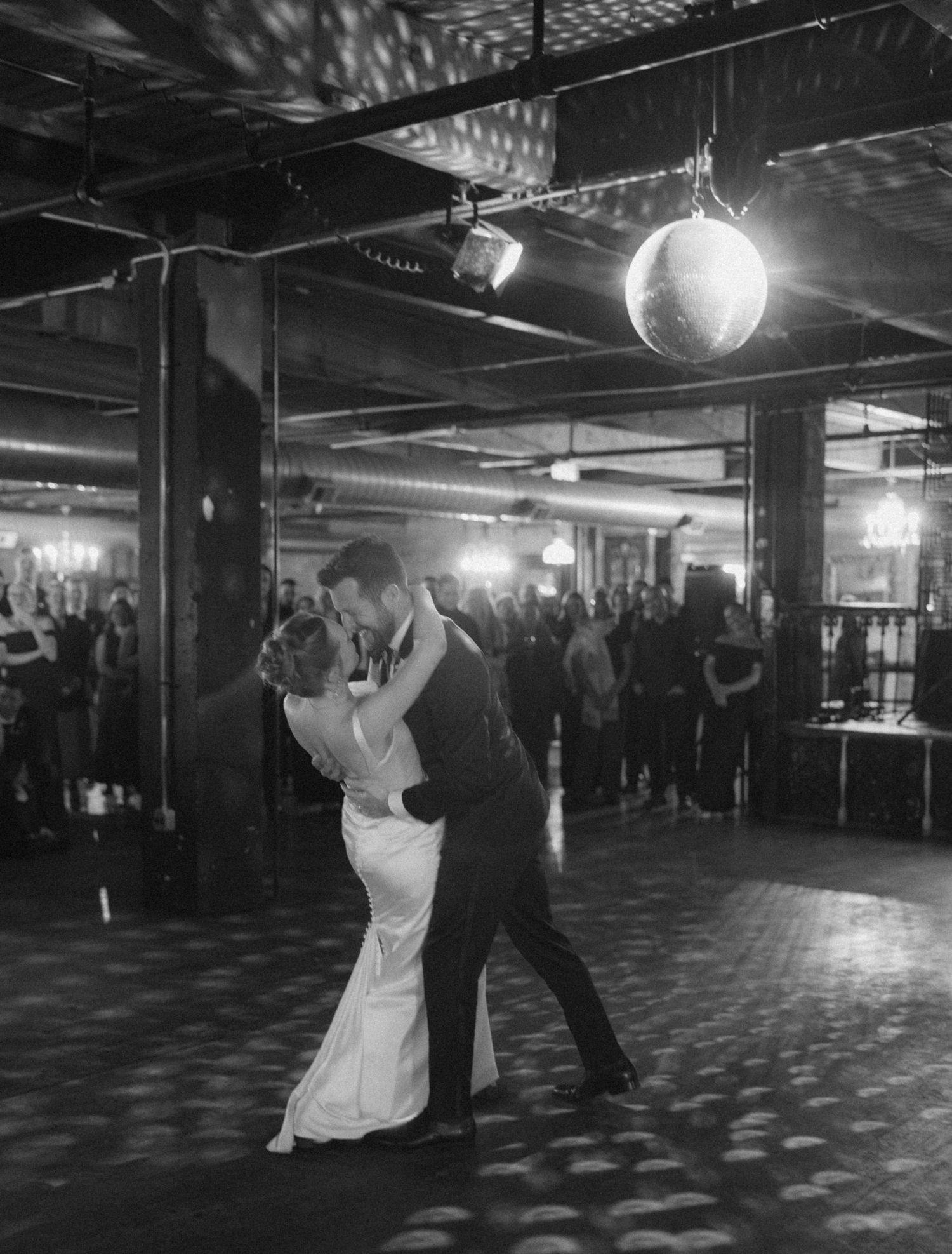 A couple shares their first dance under a disco ball at a wedding reception in a dimly lit industrial venue.