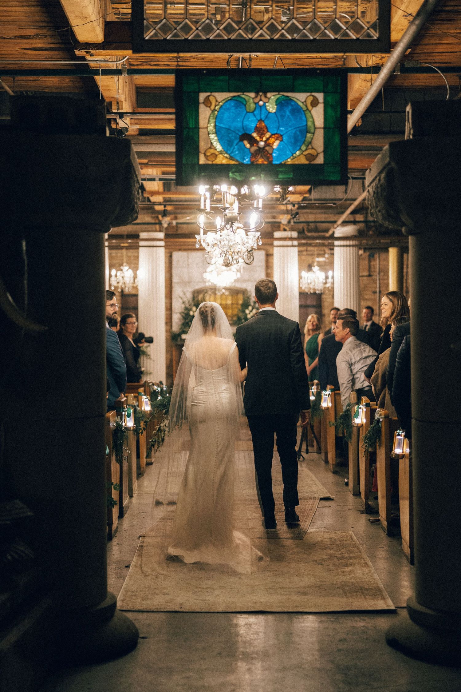 A wedding ceremony procession in a grand historic church with ornate columns and chandeliers illuminating the aisle.