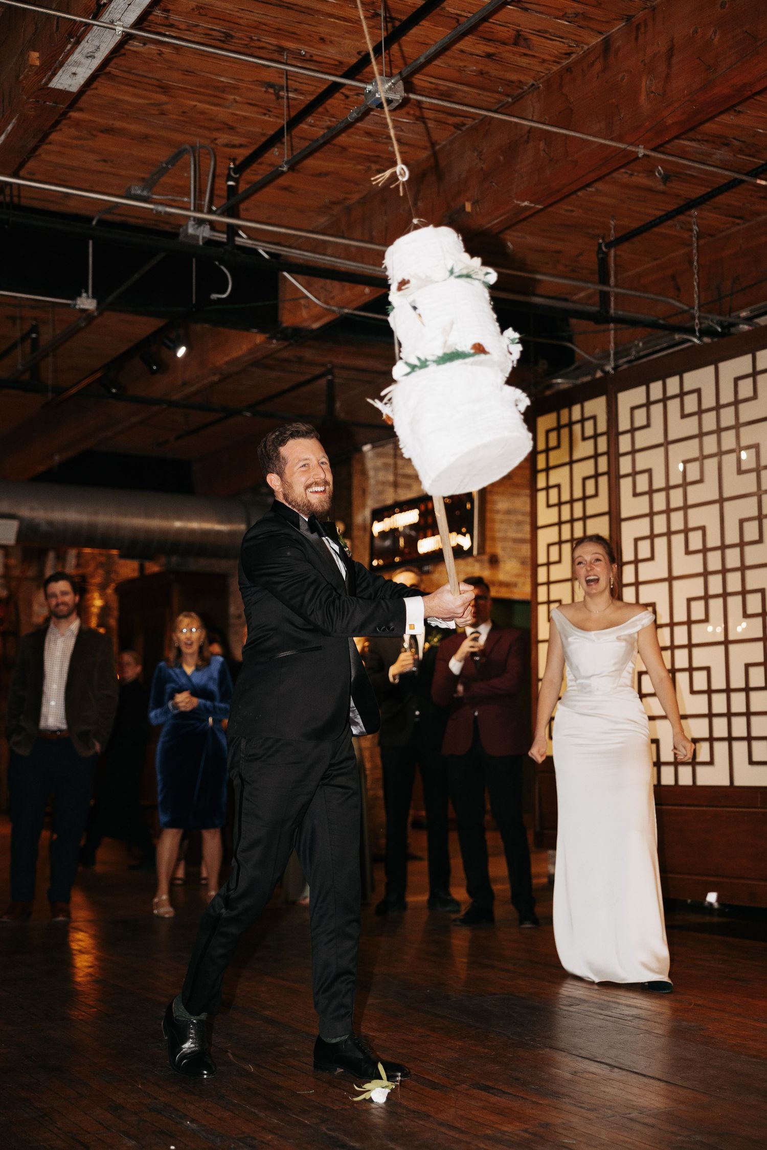 A wedding celebration with a groom balancing a tall white wedding cake while guests watch at an indoor venue with wooden floors.