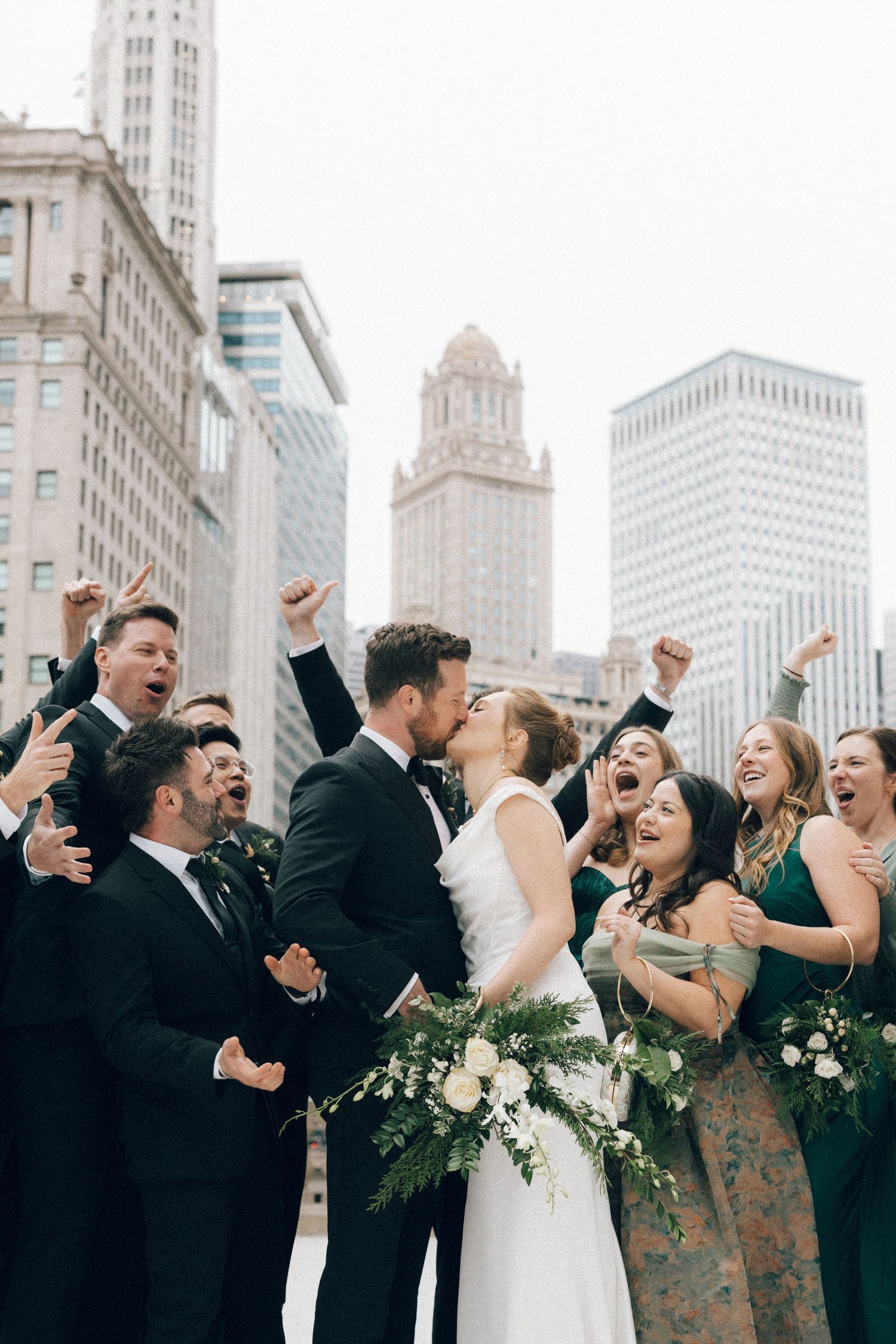 Wedding party celebrates joyfully with raised arms against urban backdrop of tall buildings and skyscrapers.