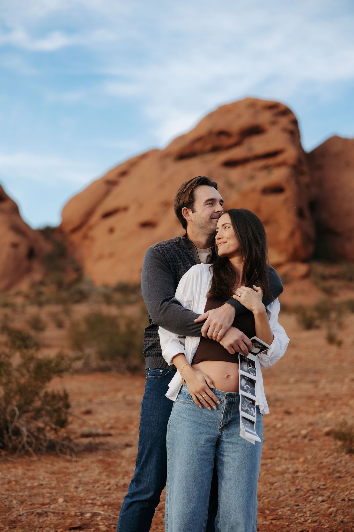 Couple embracing in red rock desert landscape, woman holding ultrasound photos, maternity announcement photo session.